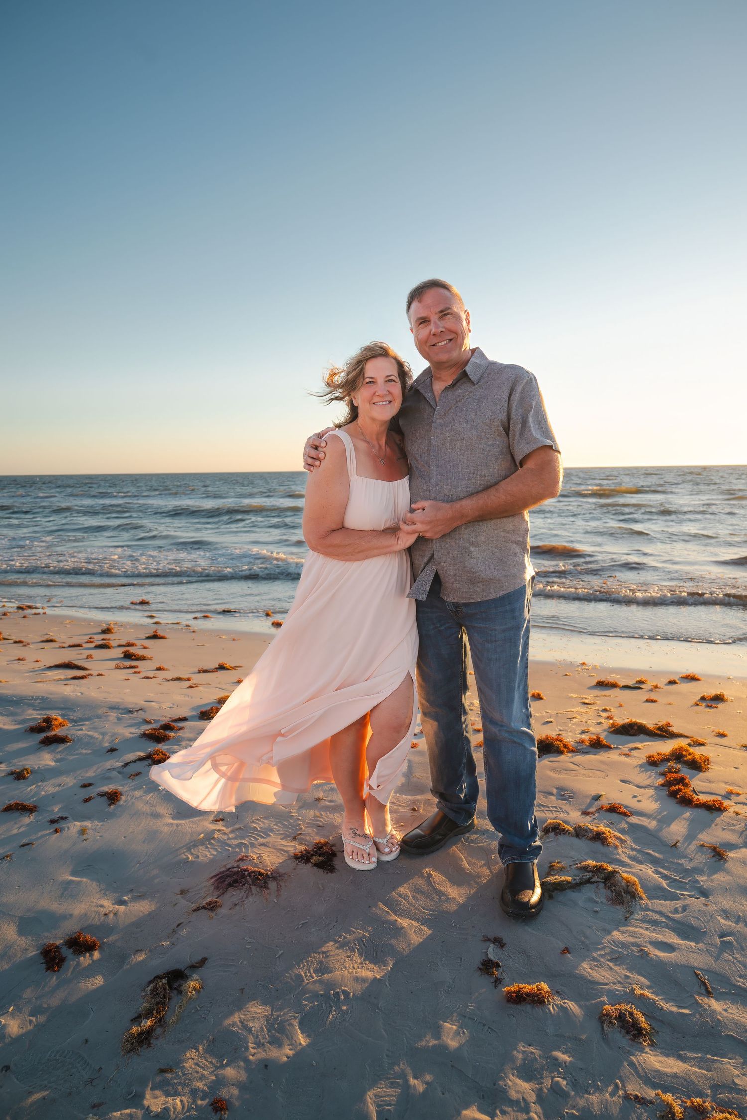 Couple embracing on a sandy beach at sunset, woman in pink dress, man in gray shirt and jeans.