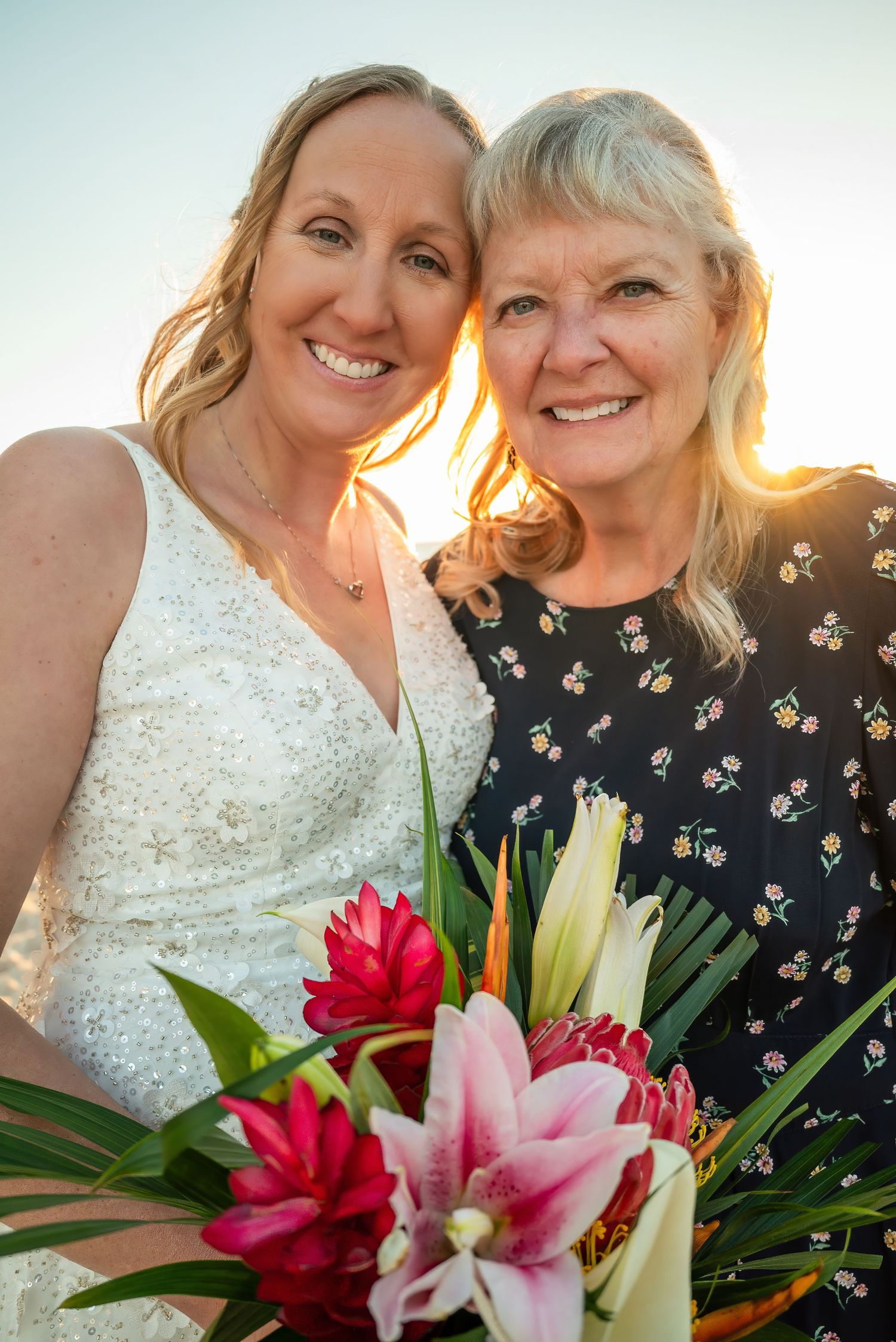 Bride and older woman smiling together at sunset beach wedding holding tropical bouquet.