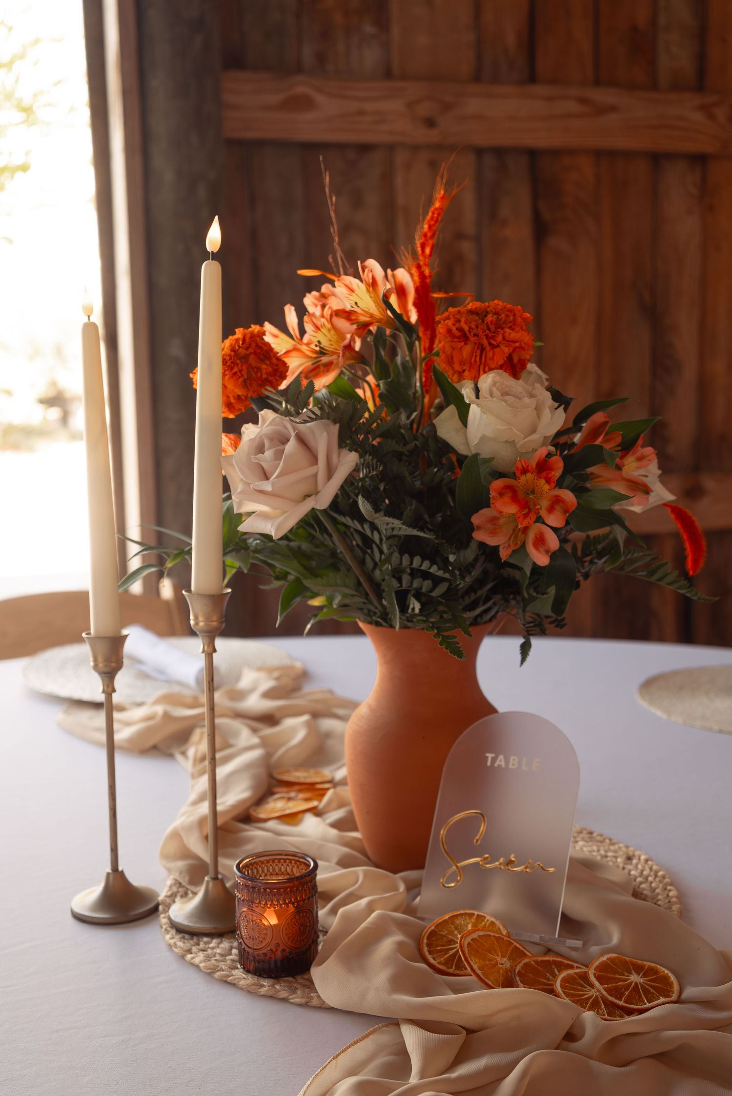 Elegant fall wedding centerpiece with orange roses, alstroemeria, candles, dried citrus, and terracotta vase on table.
