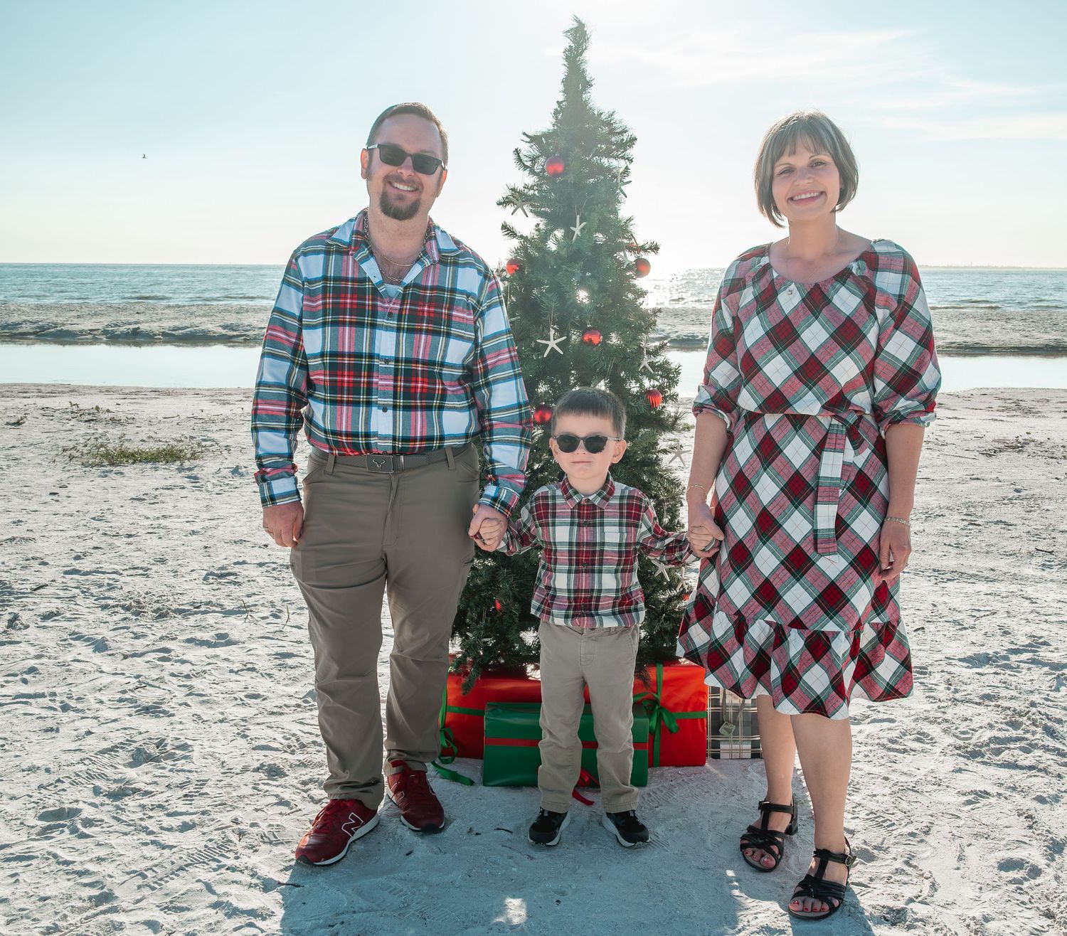 Family of three in matching plaid outfits poses beside small decorated Christmas tree on sunny beach holiday photo.