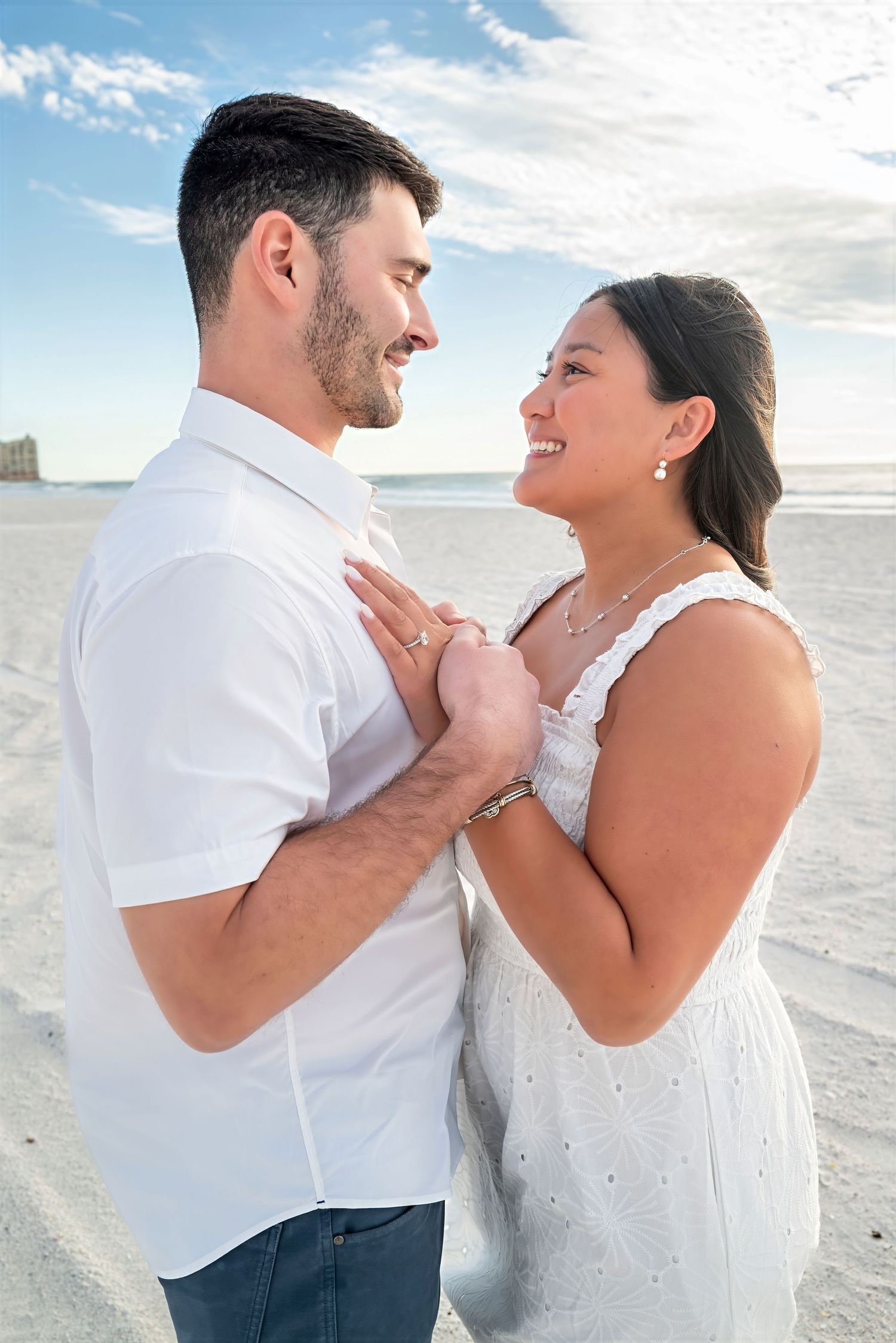 Smiling couple in white outfits embracing on sandy beach.
