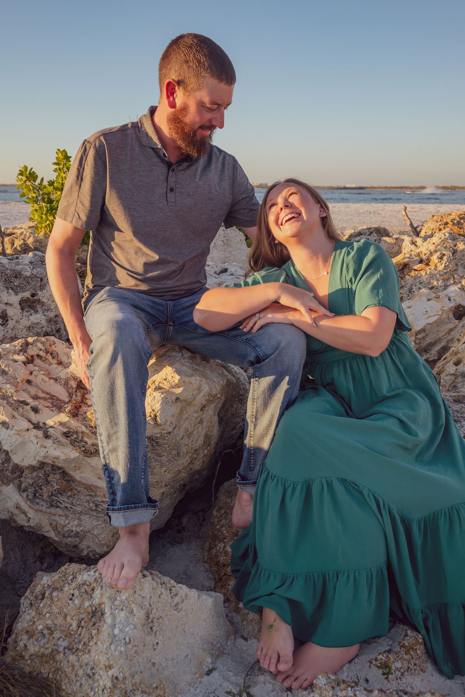 Couple laughing and sitting on rocky beach at golden hour sunset.