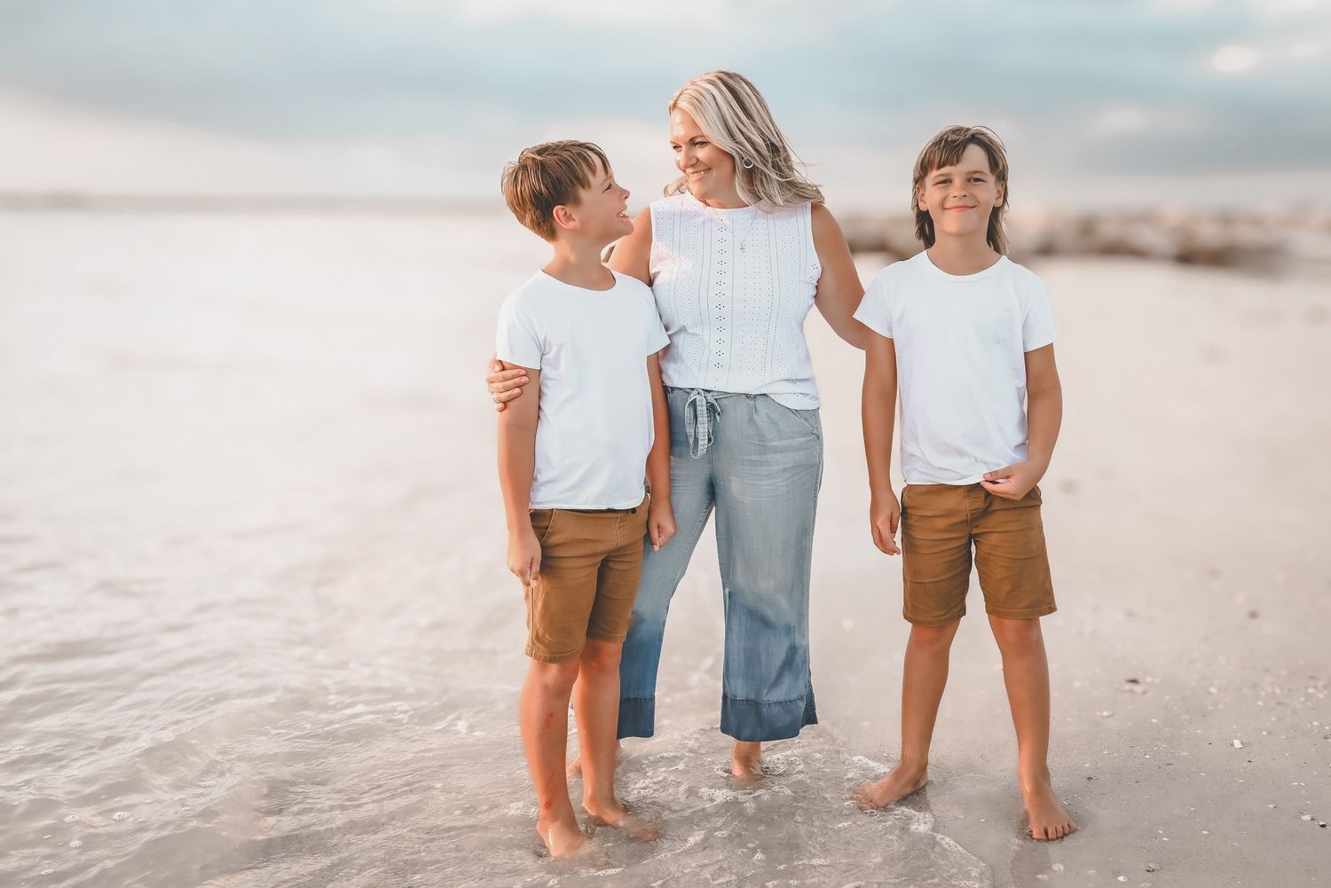 Smiling mother standing between her two young sons wearing white shirts and brown shorts on a sunny beach.