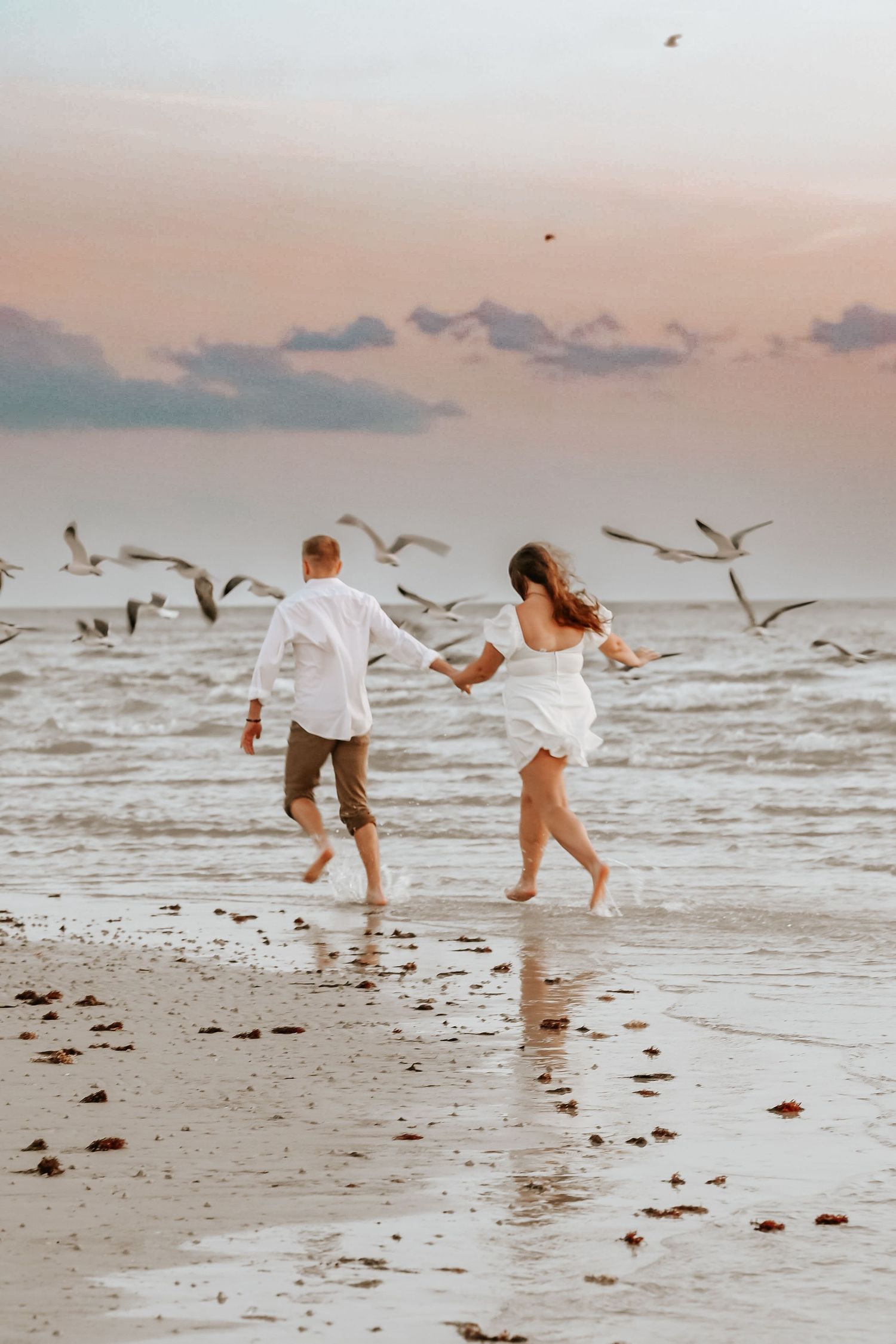 Couple running barefoot along shoreline holding hands as seagulls fly overhead at dusk.
