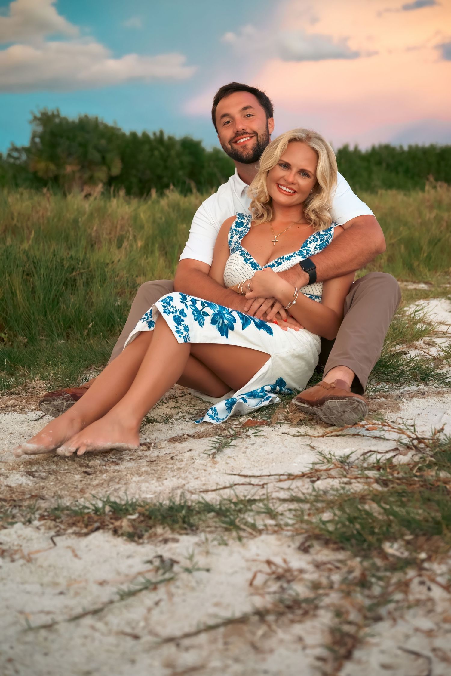 Happy couple sitting on sandy ground outdoors, woman in blue floral dress, man in white shirt, green field background.
