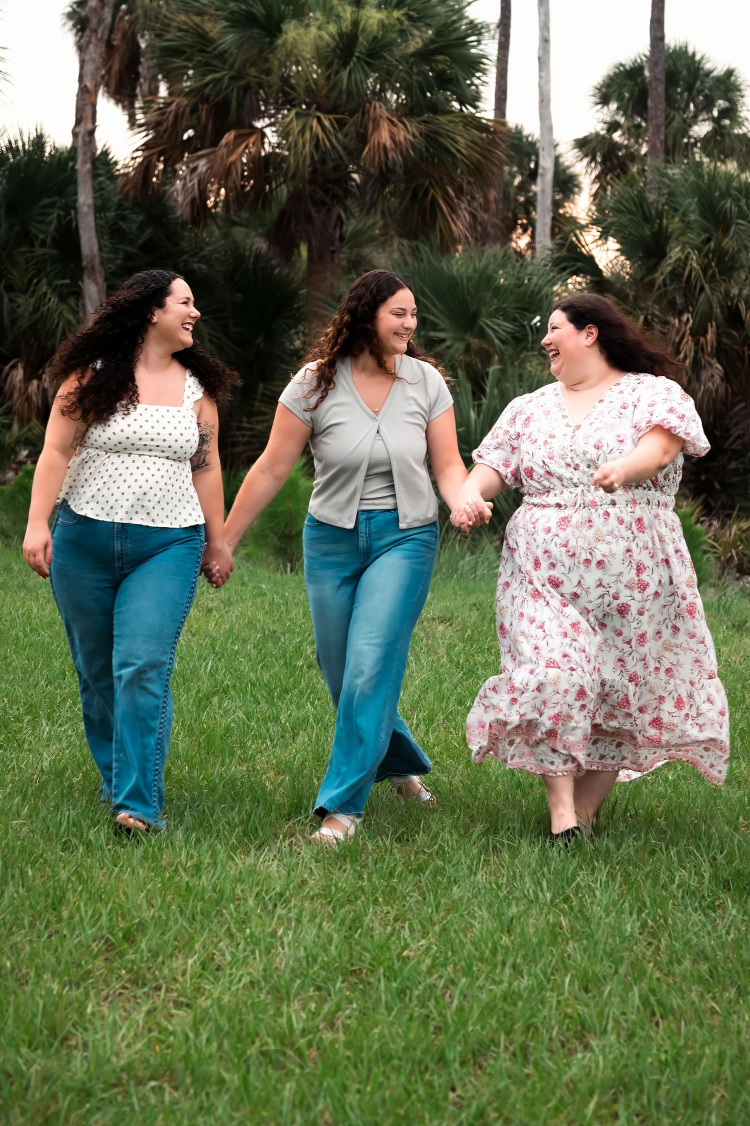 Three women holding hands and walking together on green grass with palm trees in the background.