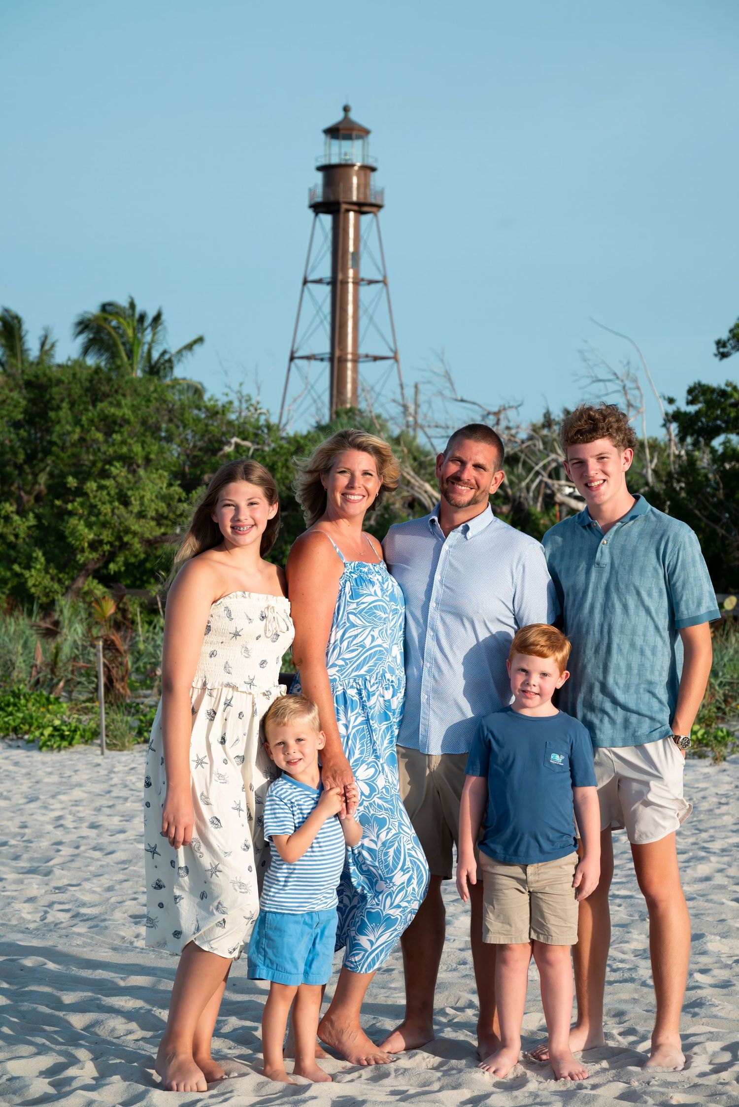 Family of six posing on white sand beach with historic lighthouse and tropical greenery in background.