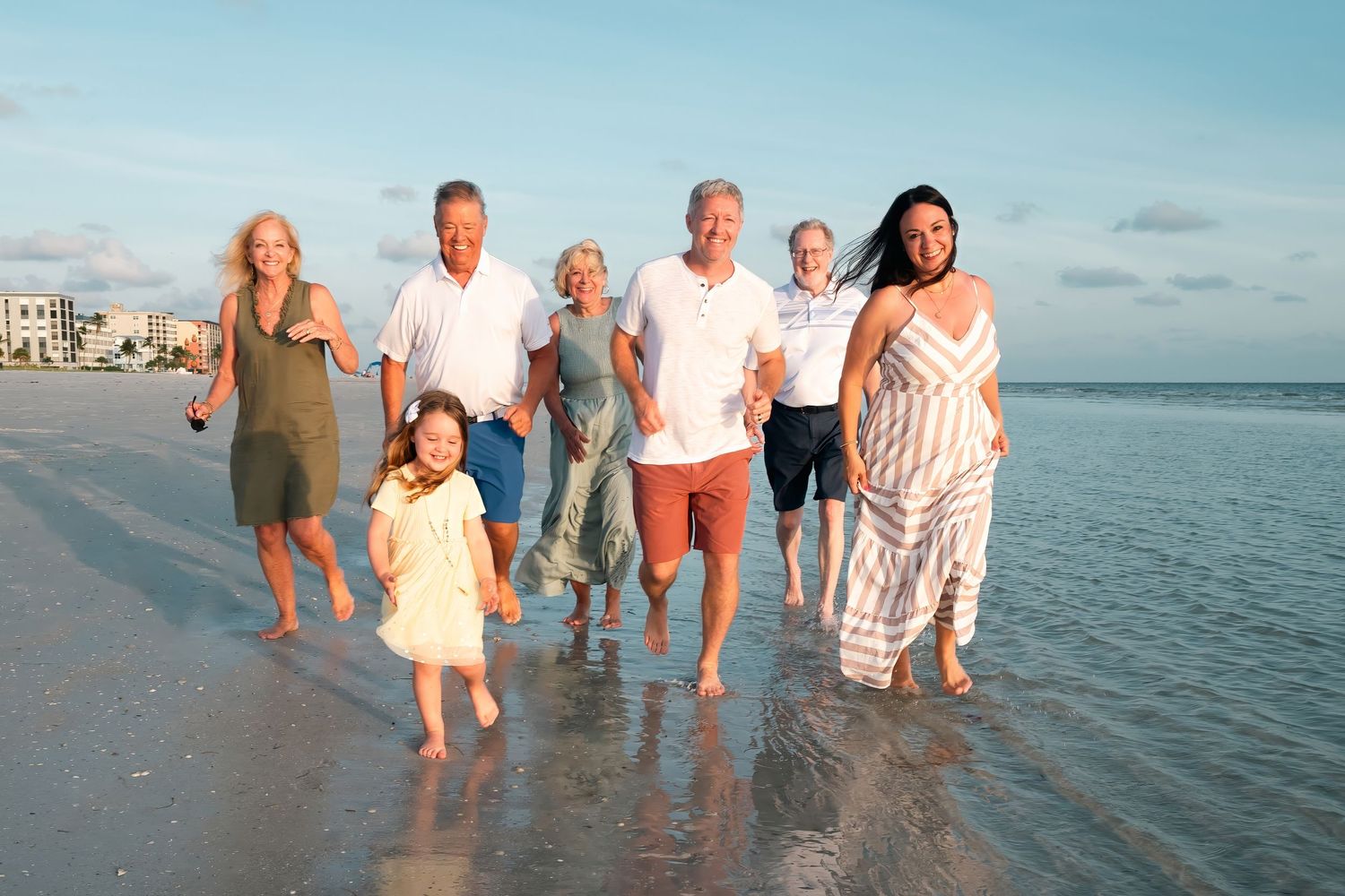 Multi-generational family smiling and walking together on a sunny beach with calm ocean waves in the background.