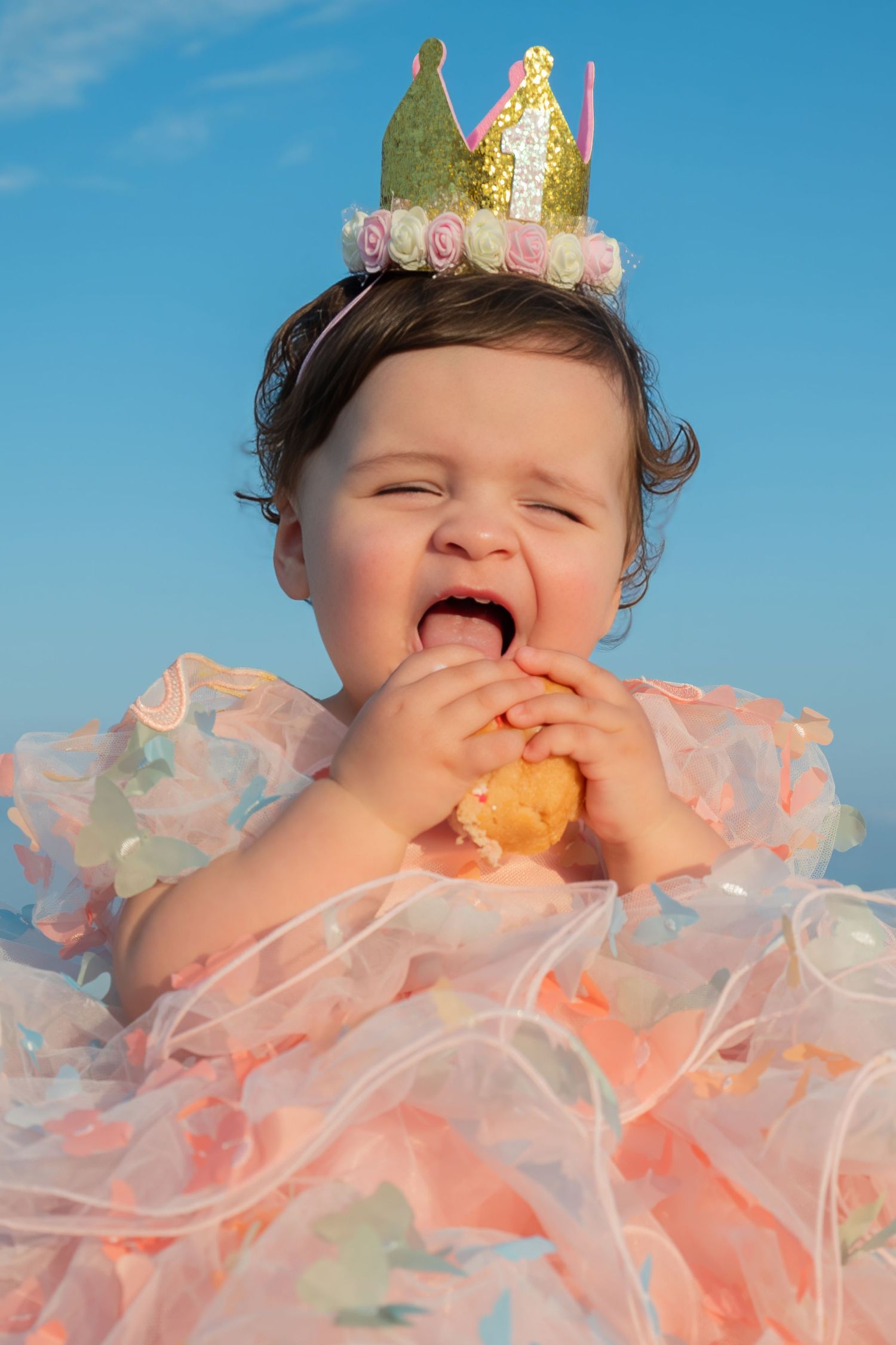 Baby girl in pink tutu dress and gold crown eating cake smash treat outdoors against blue sky.