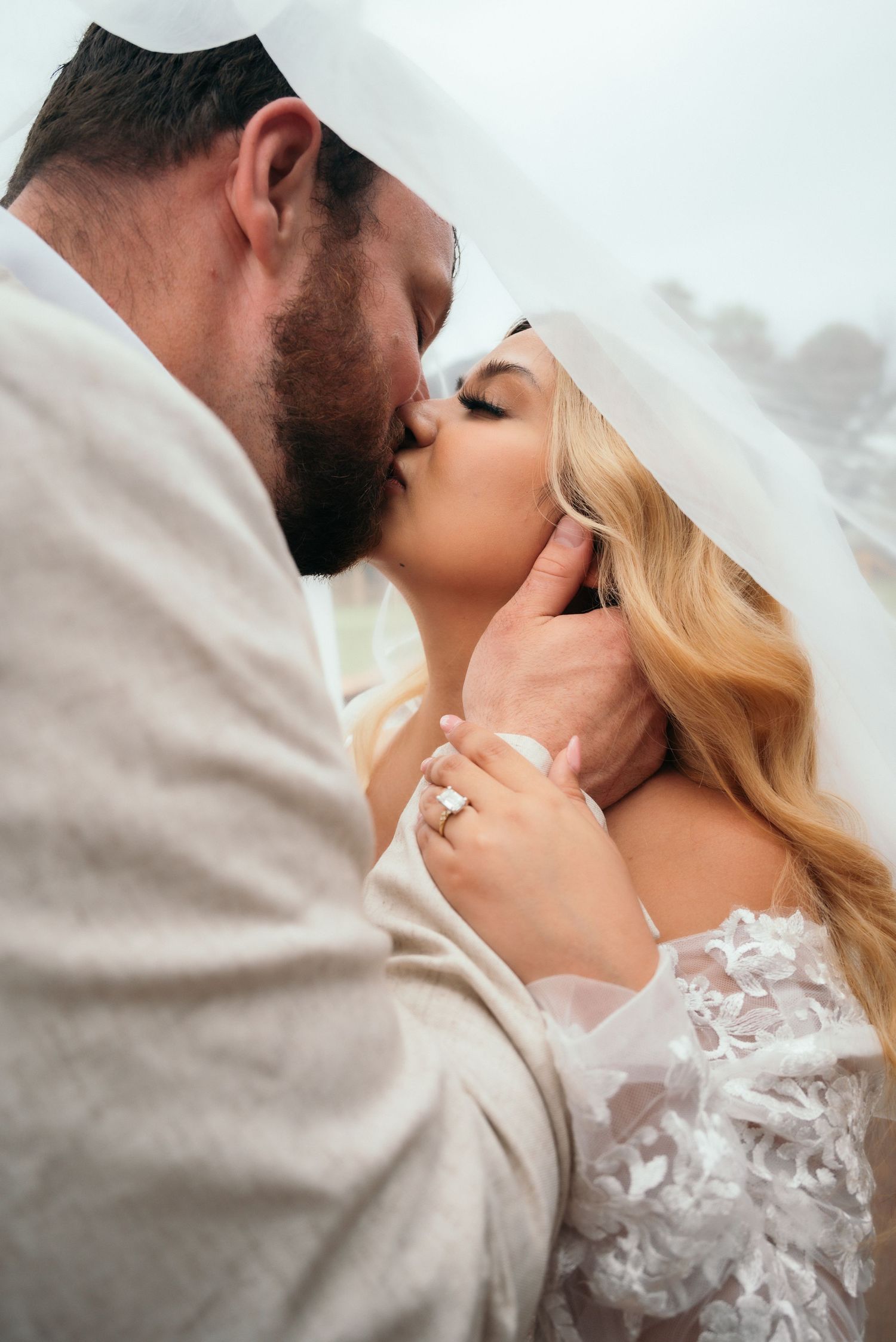 Bride and groom sharing a romantic kiss under a flowing white veil, bride wearing lace gown and diamond ring.