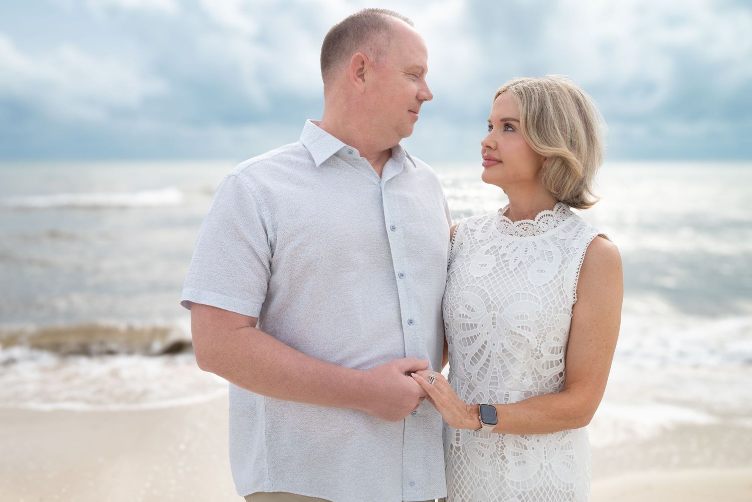 Smiling couple holding hands on beach gazing at each other with ocean waves behind them.