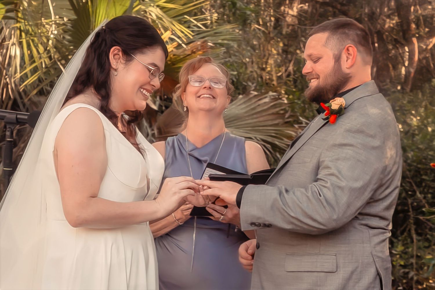 Smiling bride places ring on grooms finger as officiant watches during an intimate outdoor ceremony.