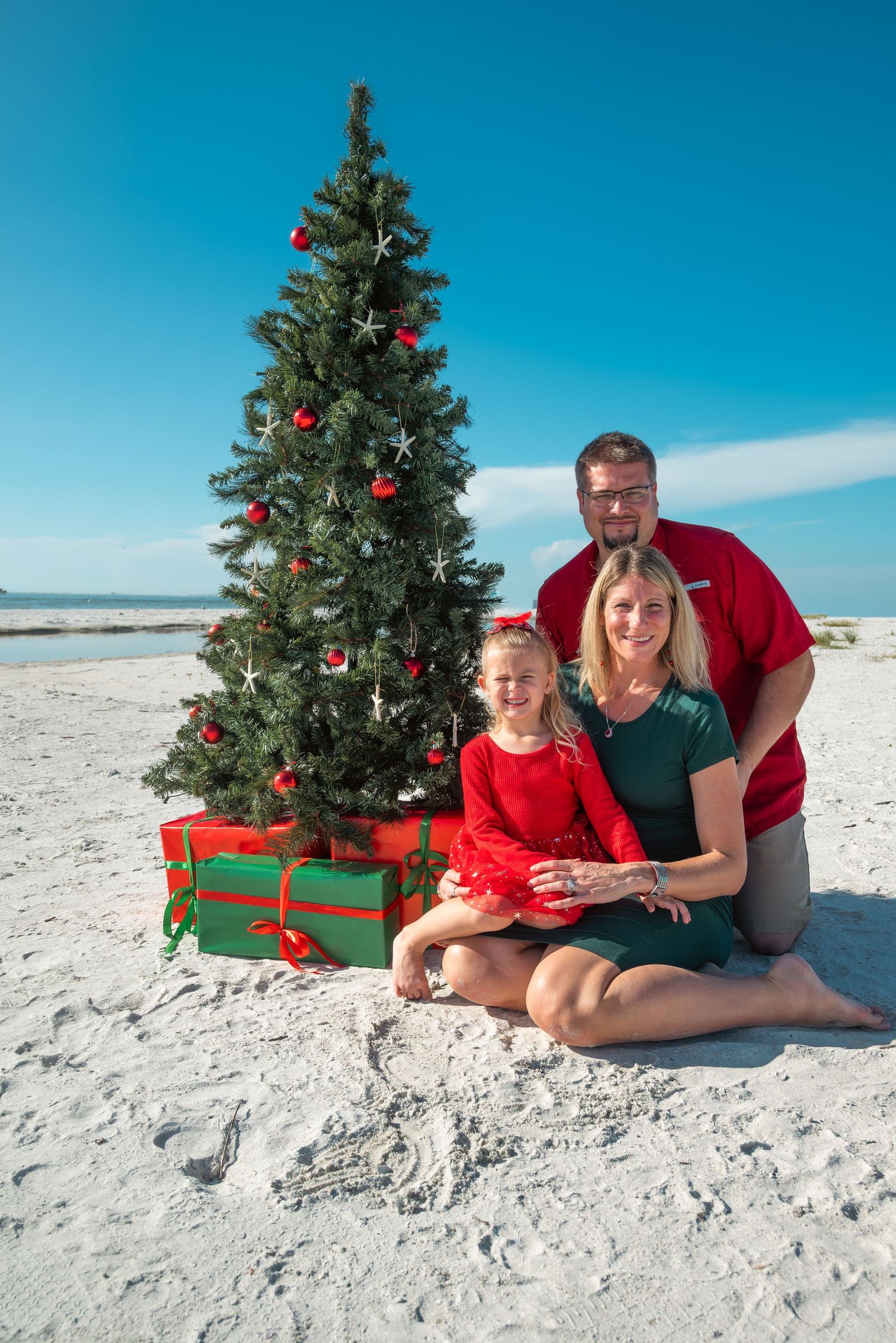 Happy family Christmas beach photo with pregnant mom in green, dad in red shirt, and young daughter by festive tree.