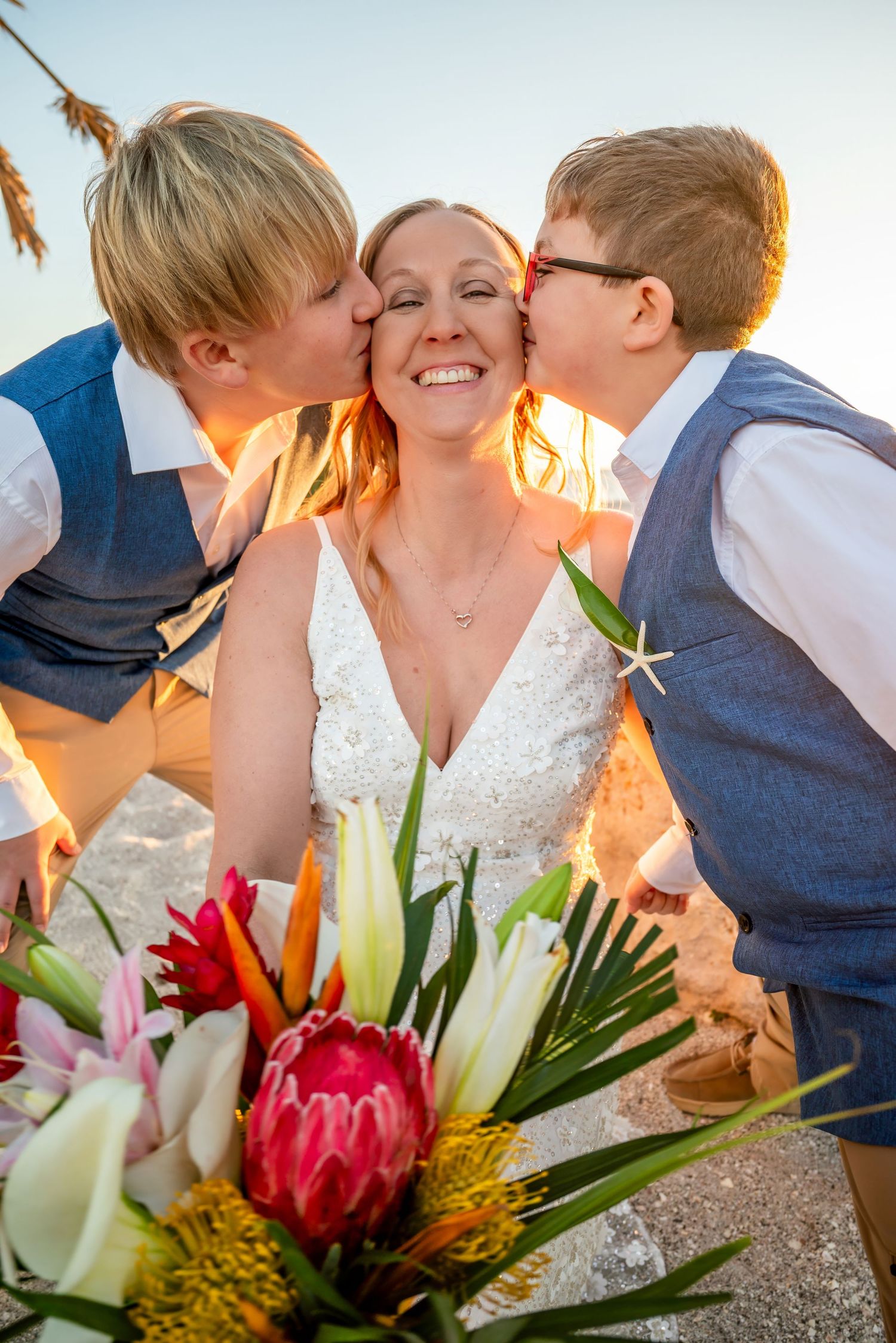 Bride smiling as two young boys in blue vests kiss her cheeks at a beach sunset wedding.