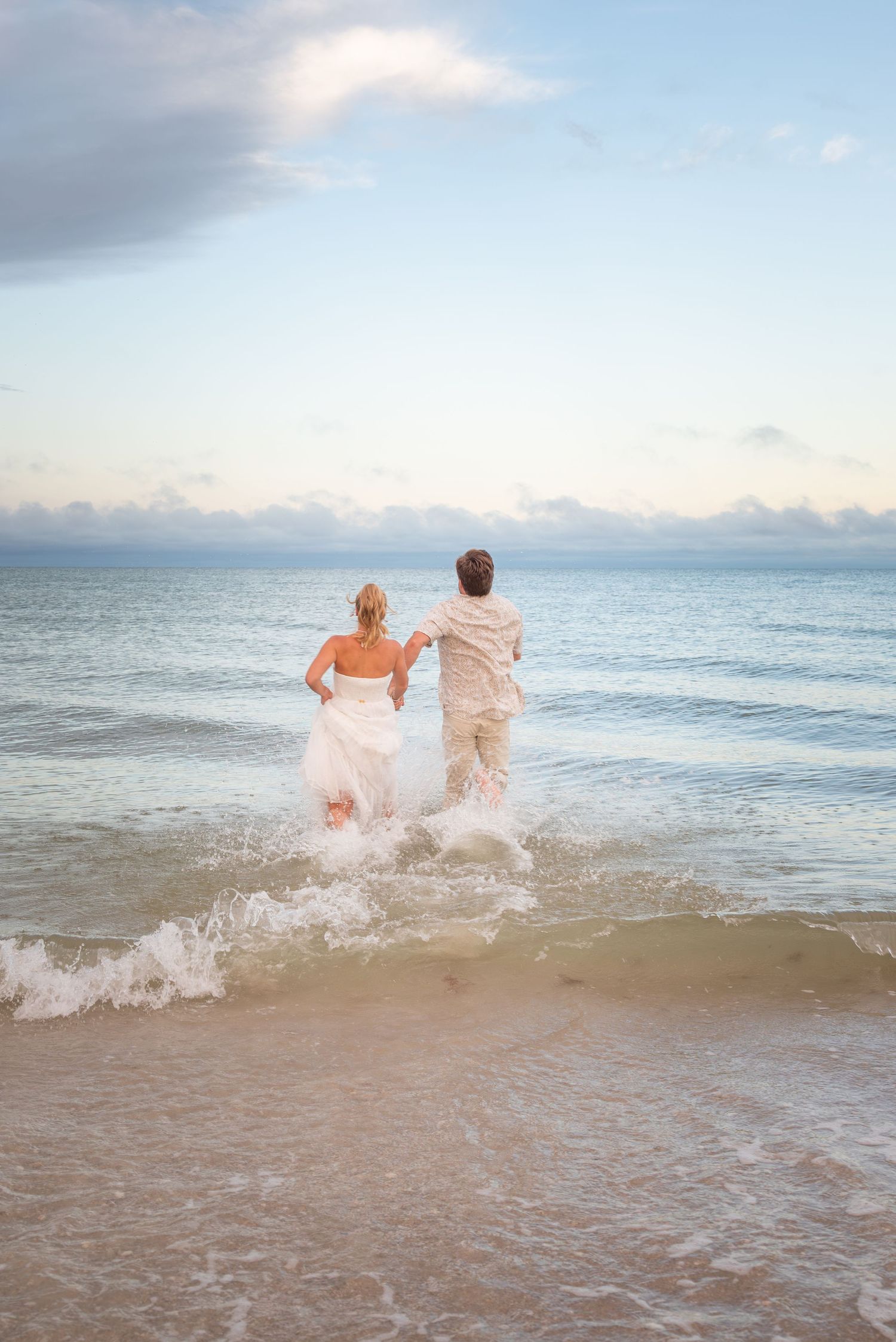 Bride and groom running into ocean waves on sandy beach in wedding attire at sunset.