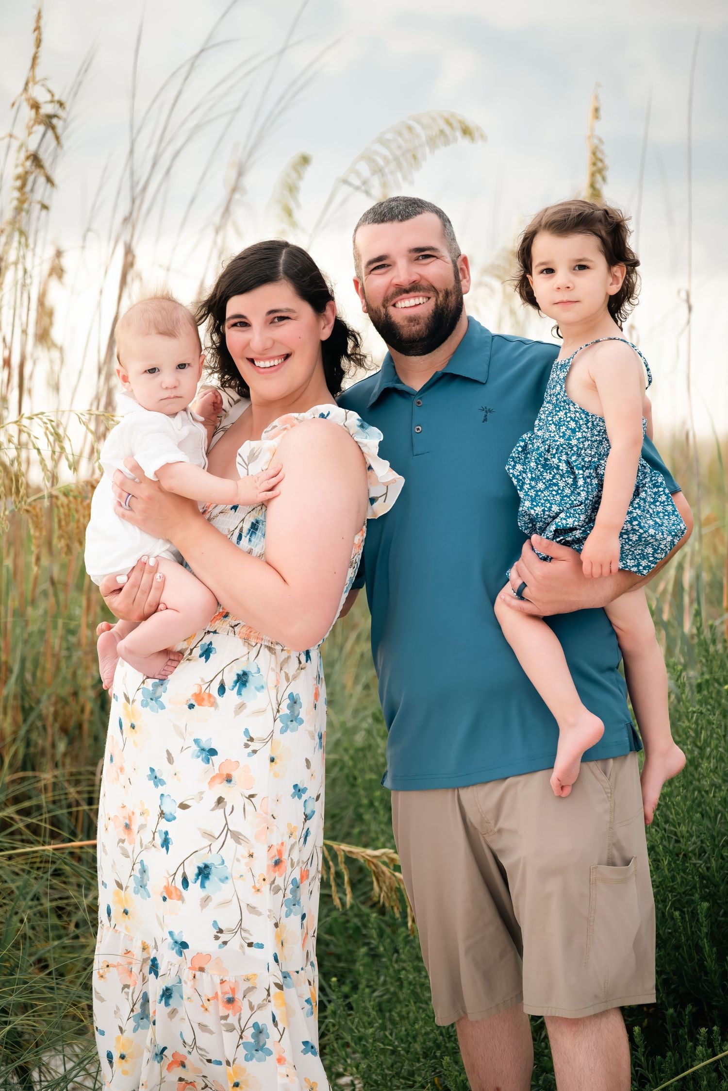 Happy family of four posing outdoors near tall sea grass, parents holding two young children, smiling at camera.