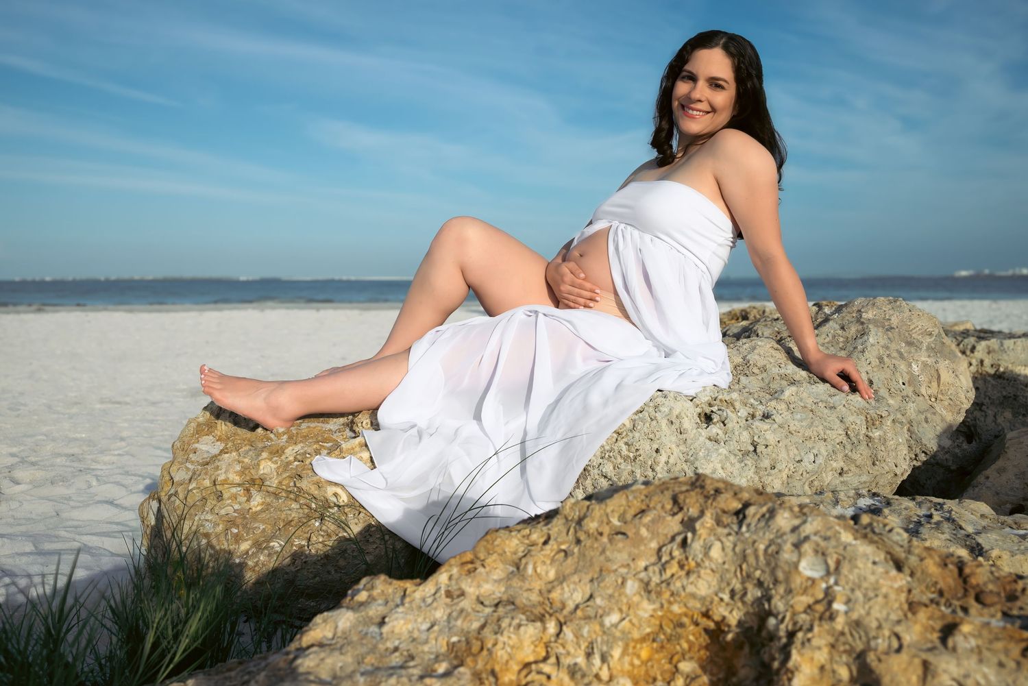 Pregnant woman in white dress sitting on rocks at a sandy beach, smiling under a blue sky.