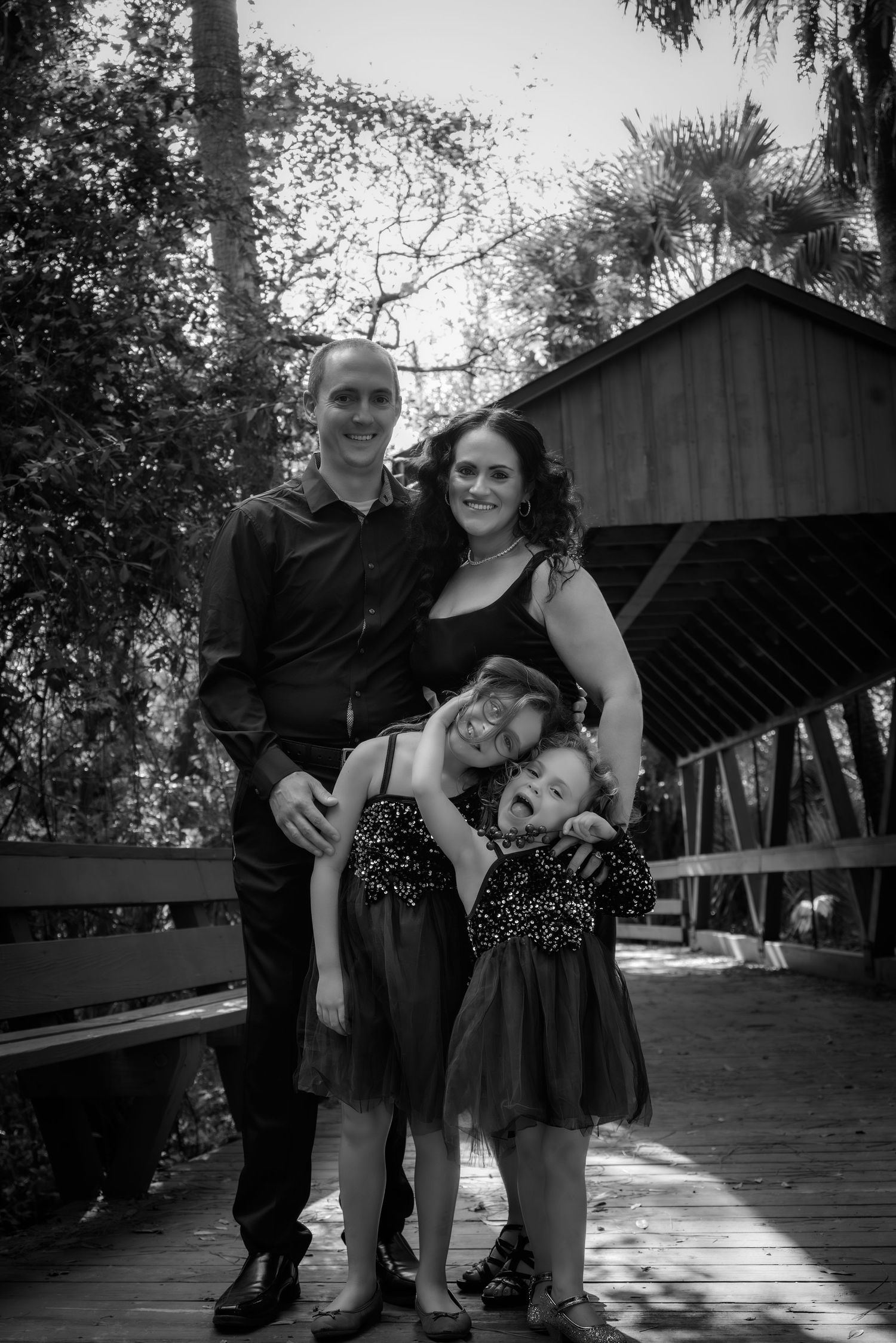 Black and white portrait of a happy family of four posing on a wooden bridge outdoors.