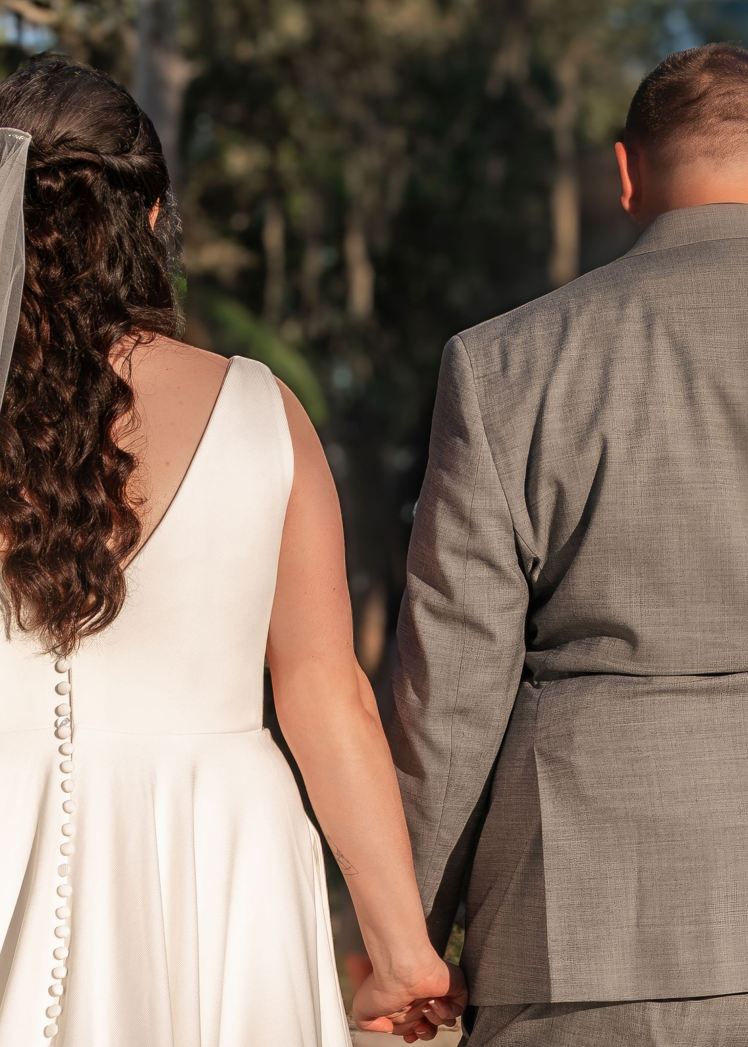 Bride in white gown and groom in gray suit hold hands while walking through a wooded setting.