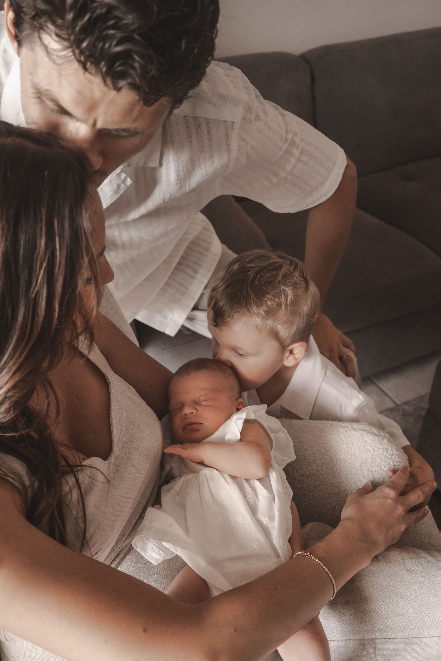 Family of four gathers closely as toddler kisses sleeping newborn held by mother.