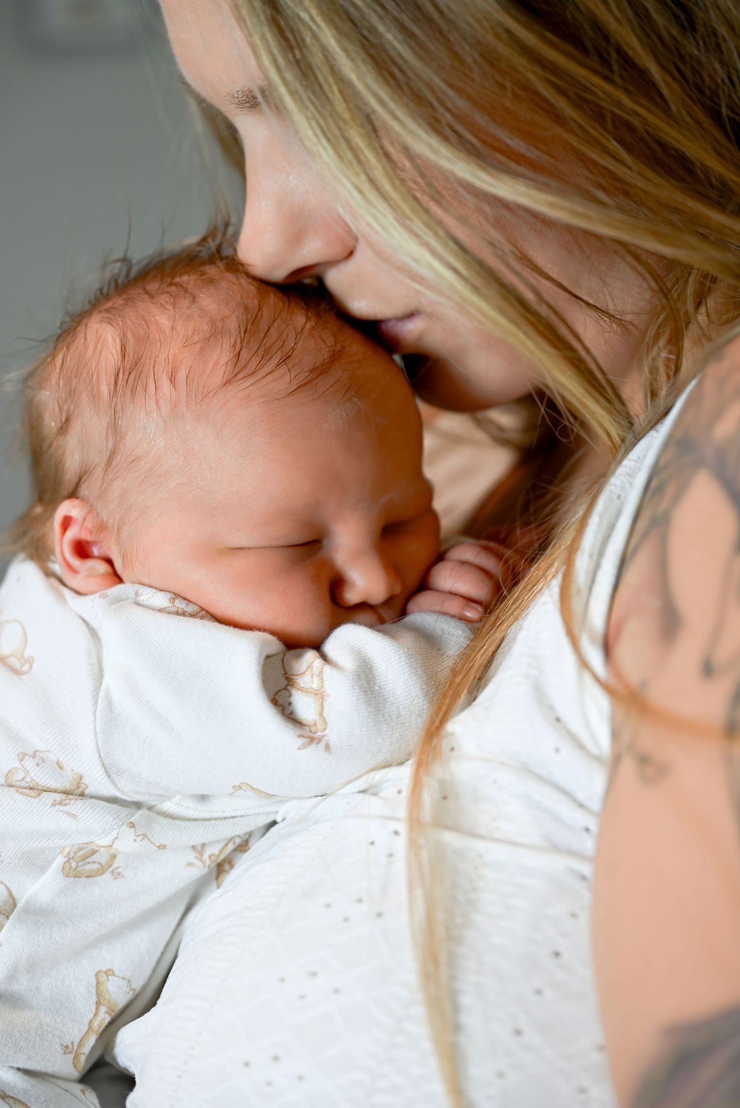 A blonde mother with tattoos gently kisses her sleeping newborn baby wrapped in a white swaddle blanket.