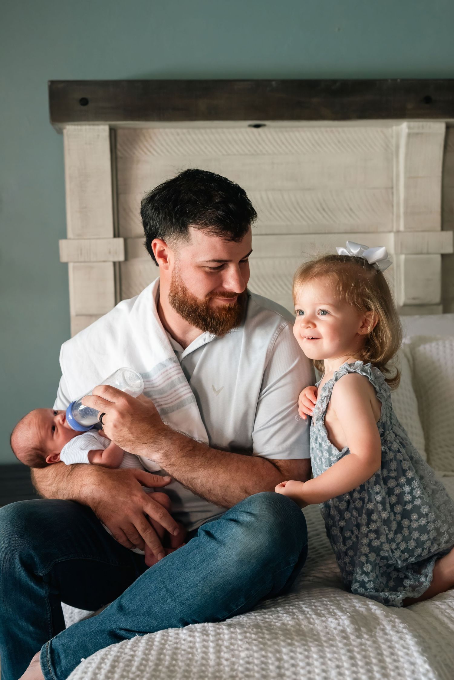 Bearded father sits on bed holding newborn baby while toddler girl in blue dress smiles beside him.