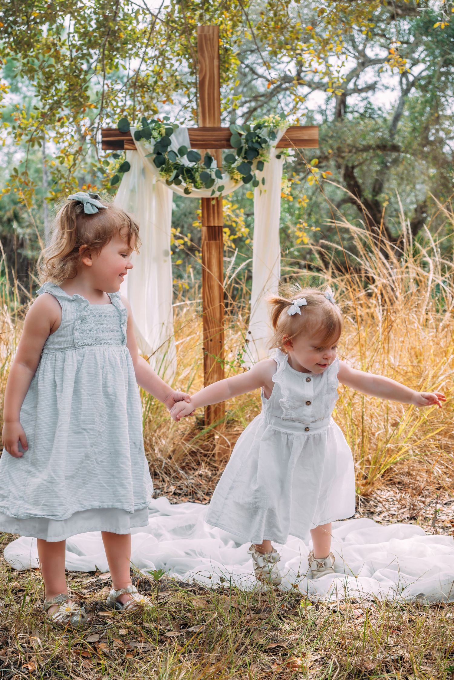 Two little girls in light blue dresses holding hands near a wooden cross outdoors.