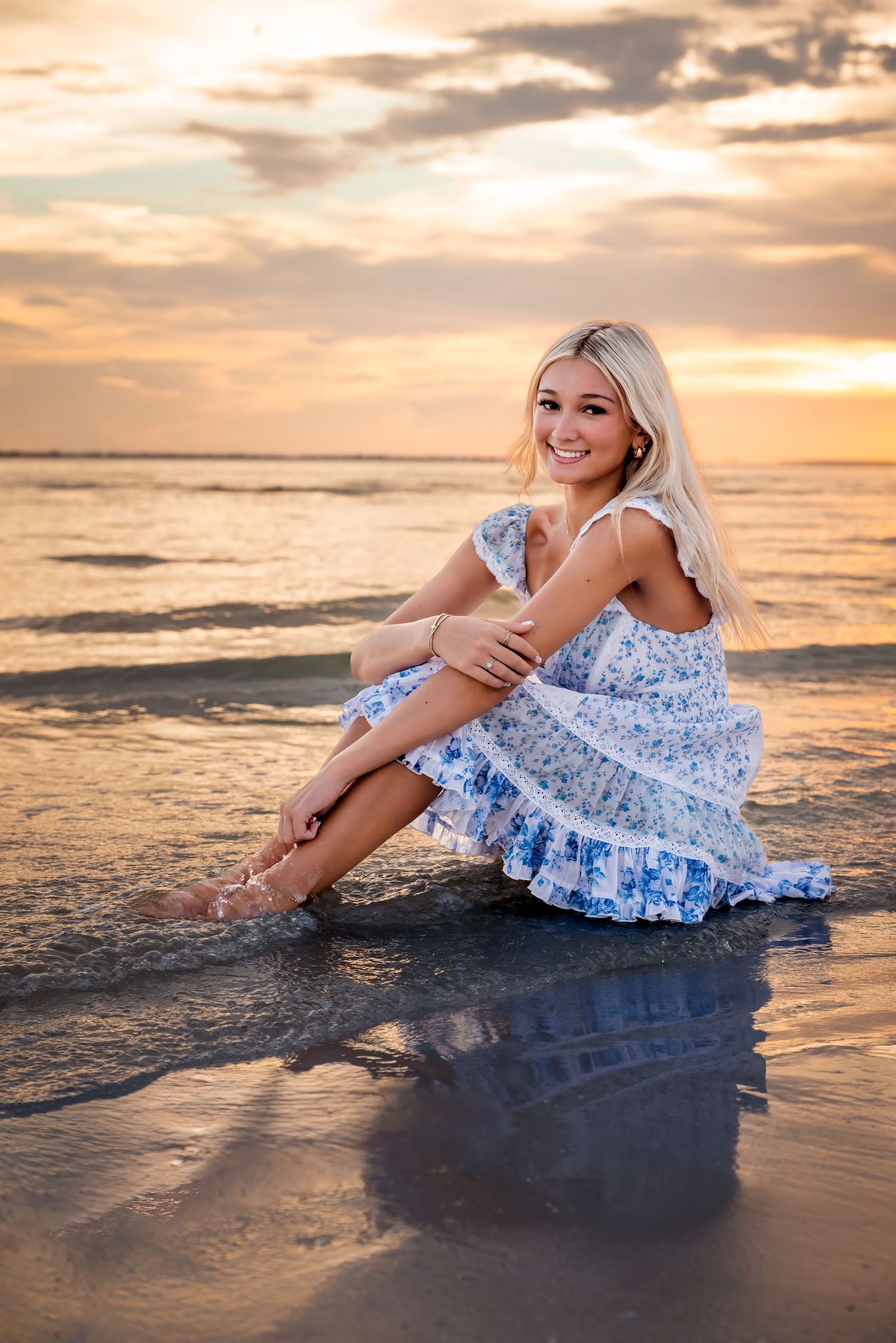 Smiling blonde woman in floral dress sitting on wet sand at beach during golden sunset.