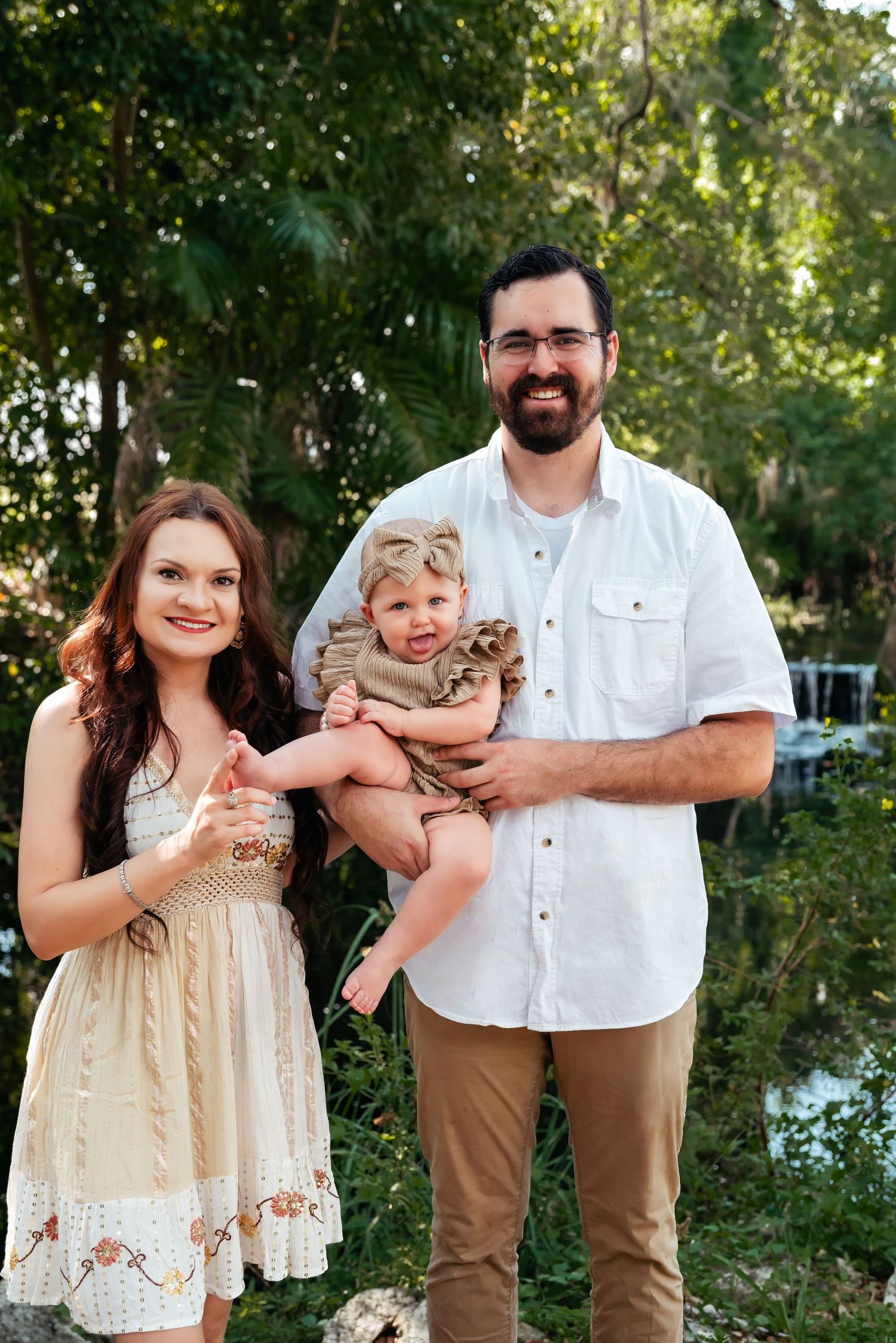 A smiling young family with a man in a white shirt, woman in a cream dress, and baby girl posing outdoors.