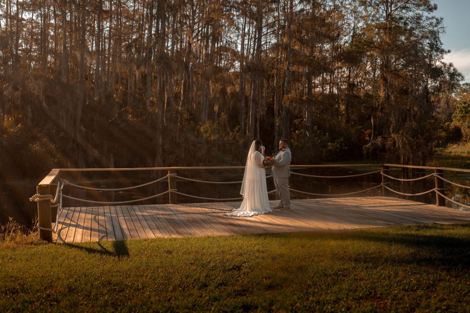 Wide-angle golden hour wedding portrait of bride and groom holding hands on a rustic wooden dock in nature.