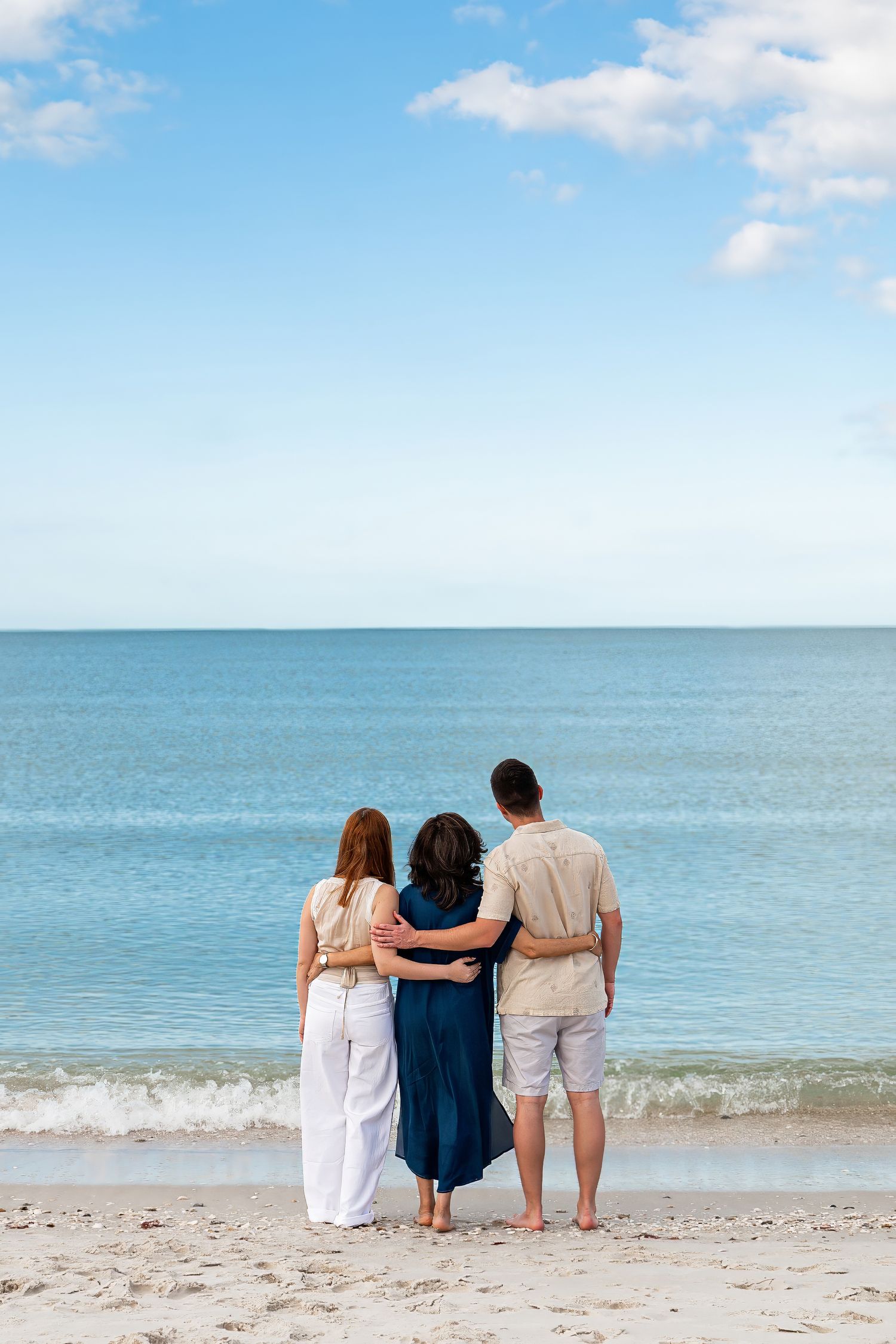 Family of three with arms around each other gazing at the ocean.
