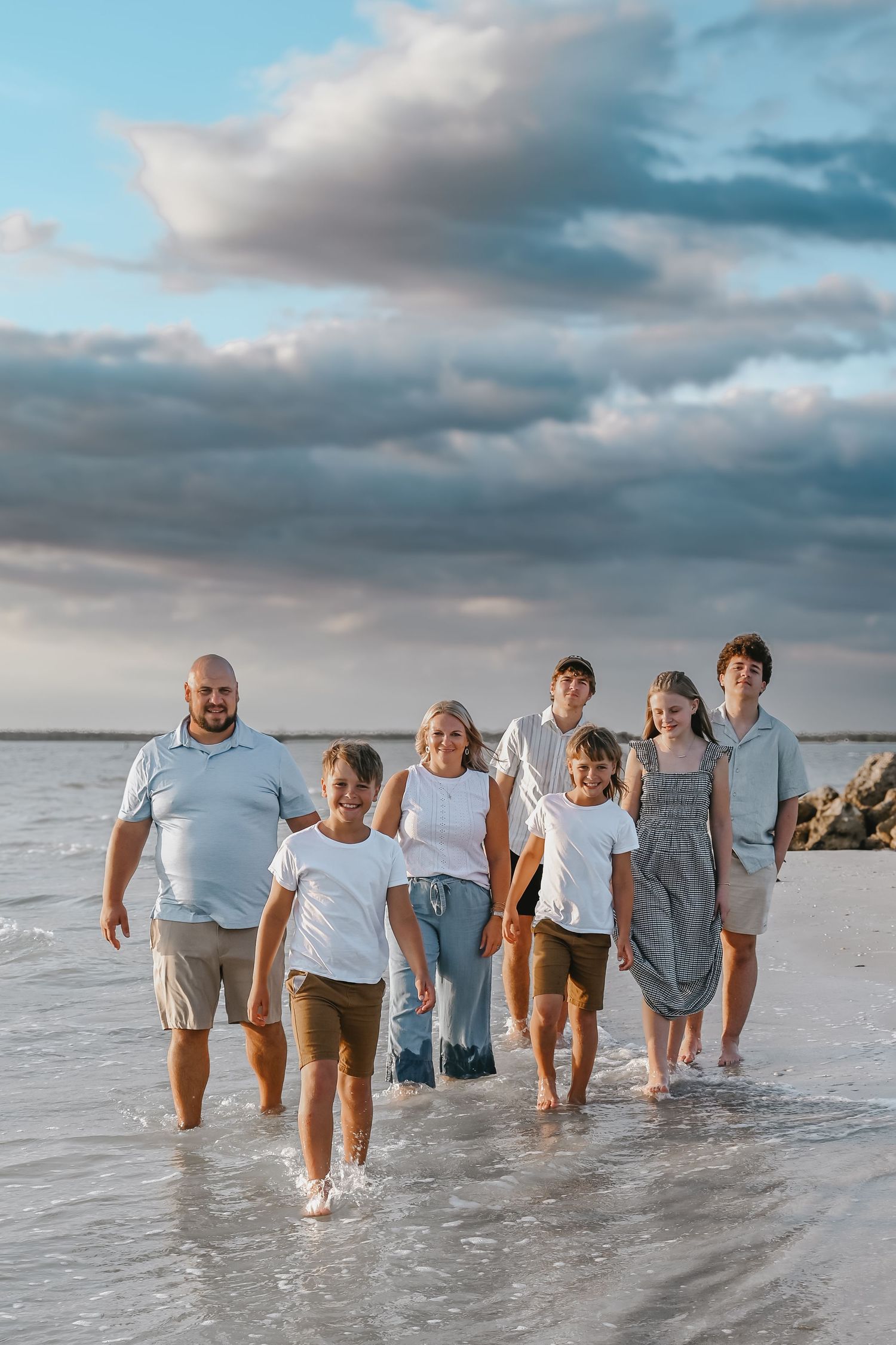 Large family of seven walking together through shallow beach water at sunset under dramatic cloudy skies.