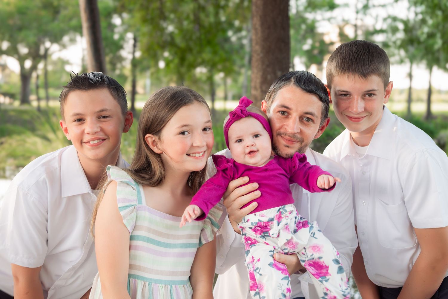 A father poses with four children including a baby in pink outdoors.