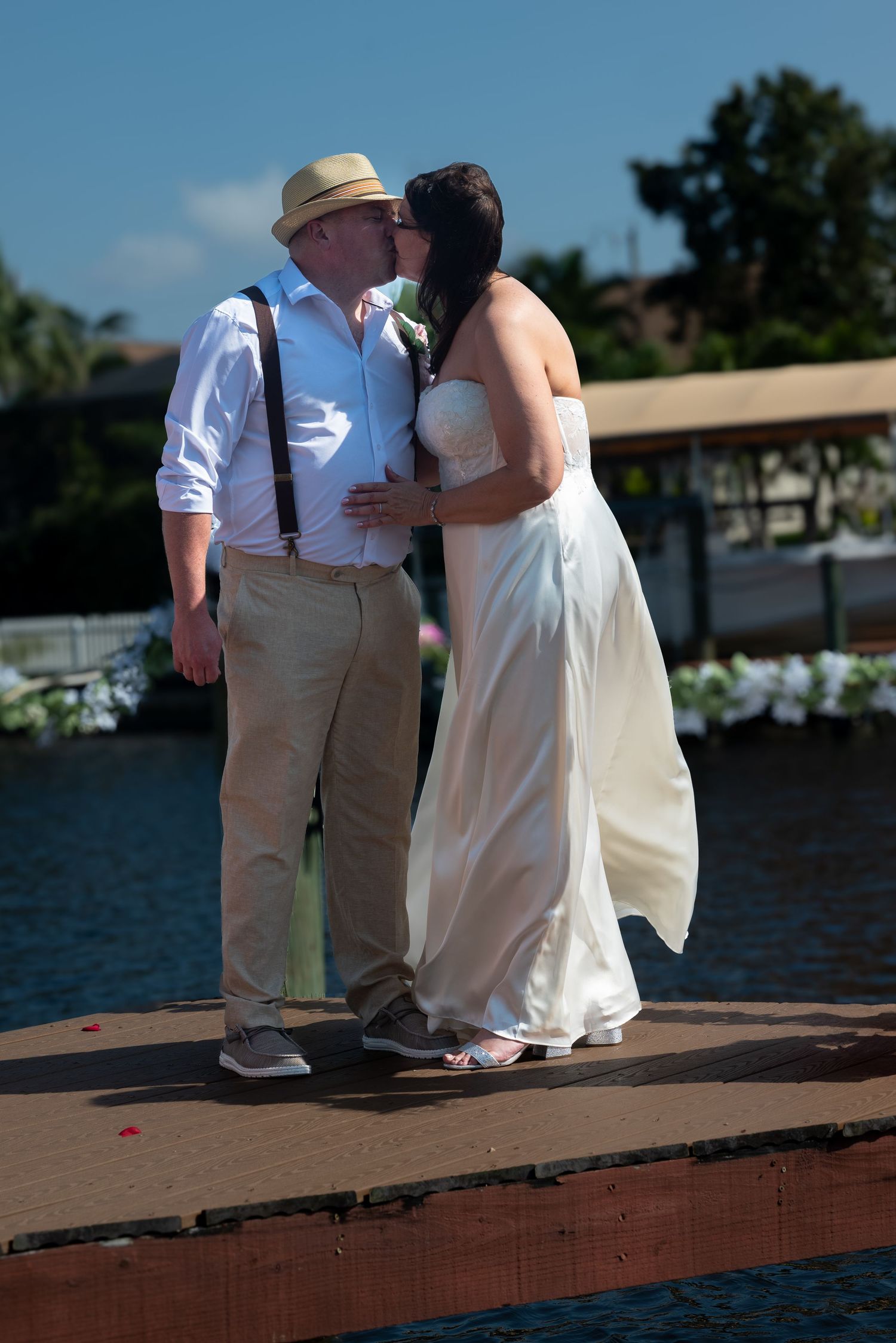 Bride and groom share first kiss on wooden dock with scenic waterway in background at outdoor wedding.