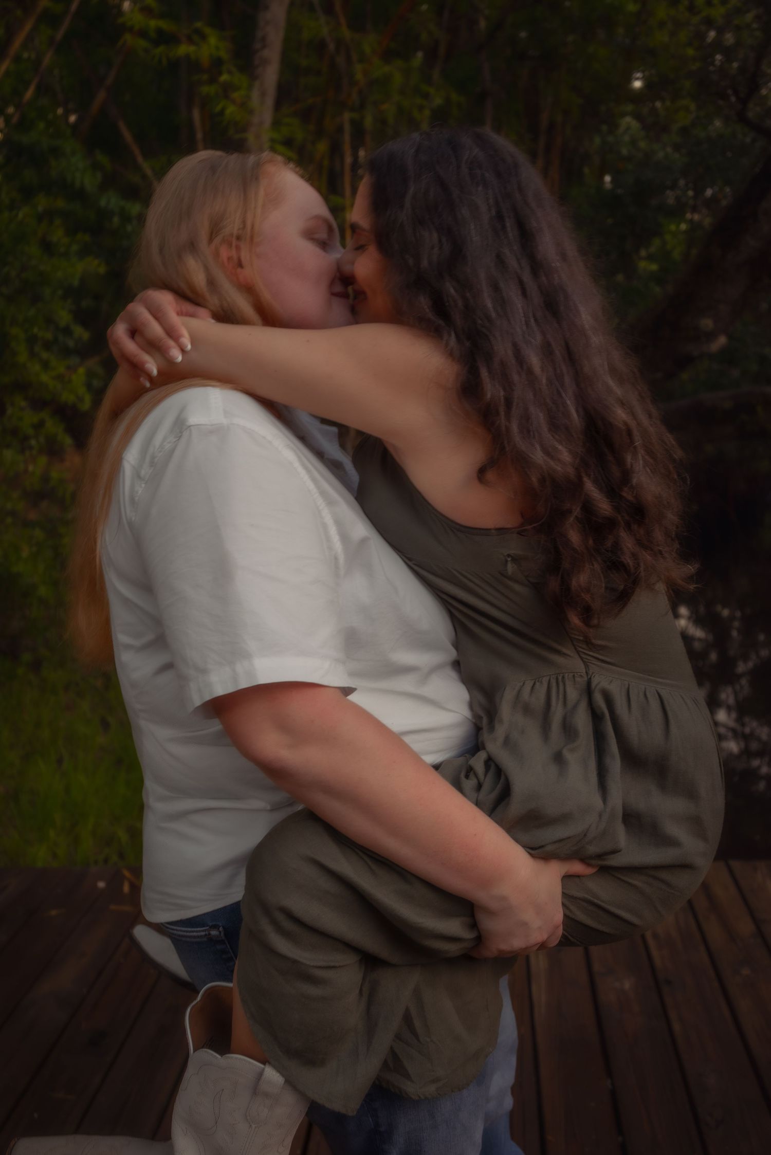 Two women share a loving kiss while embracing on an outdoor wooden deck at sunset.