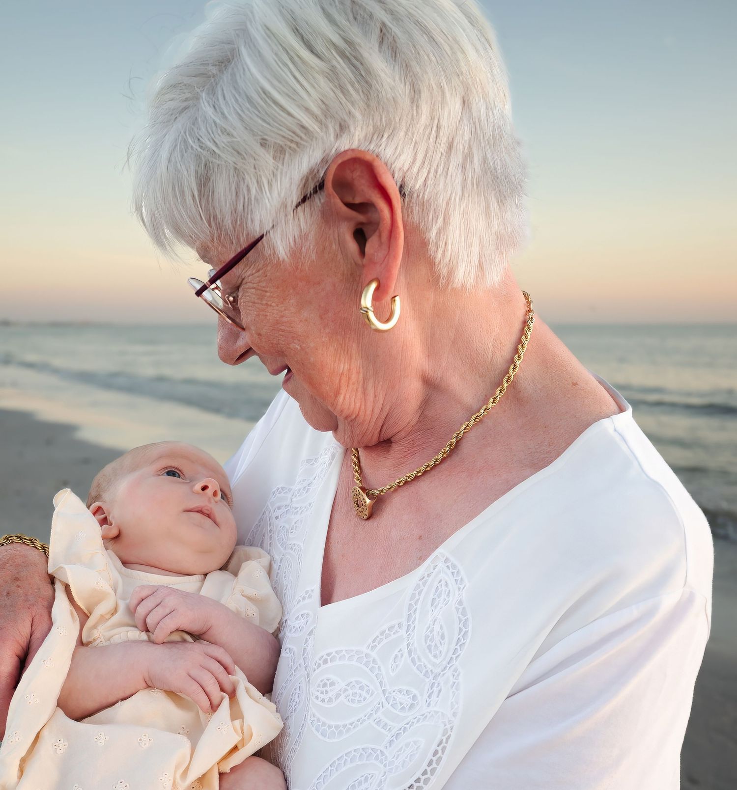 Grandmother lovingly gazing at newborn baby on a peaceful beach at sunset.
