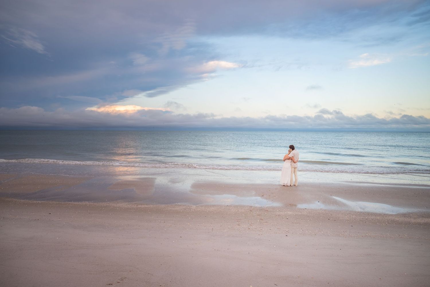 Bride in white dress standing alone on vast sandy beach at sunset with dramatic cloudy sky.