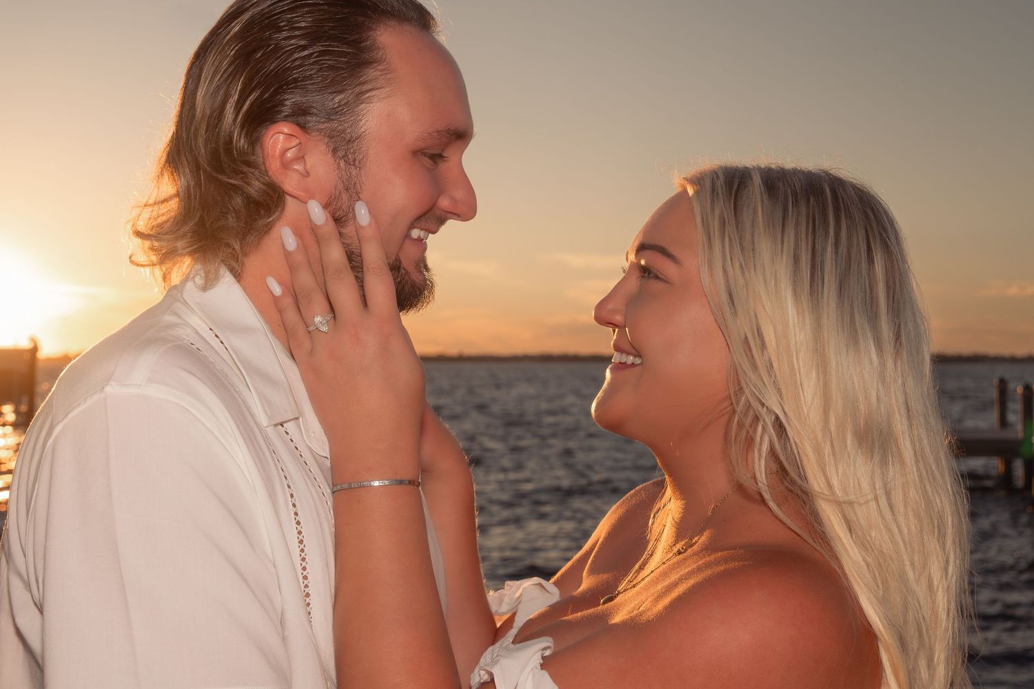 Smiling couple at sunset, woman proudly showing her engagement ring near the water.