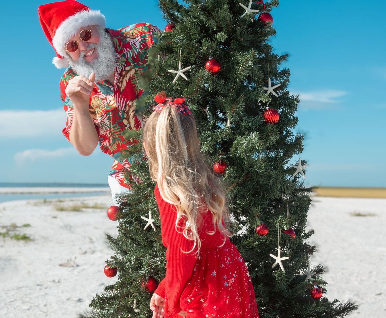 Santa in a Hawaiian shirt decorates a Christmas tree on a sunny beach with a girl in a red dress.