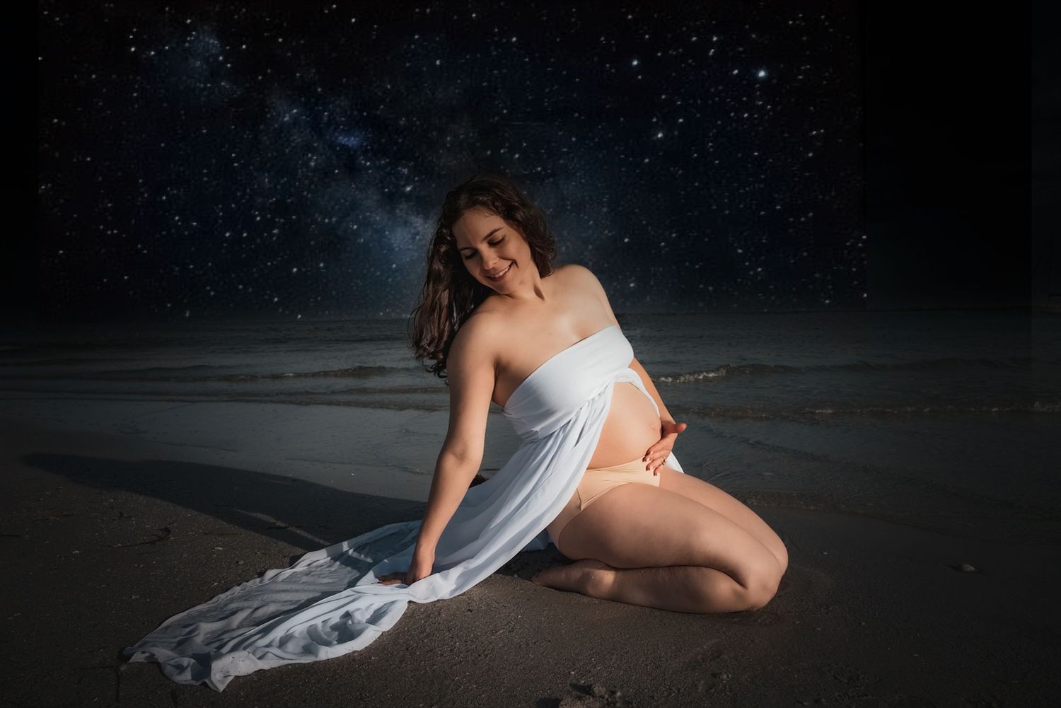 Pregnant woman in white fabric wrap poses on beach under starry night sky.