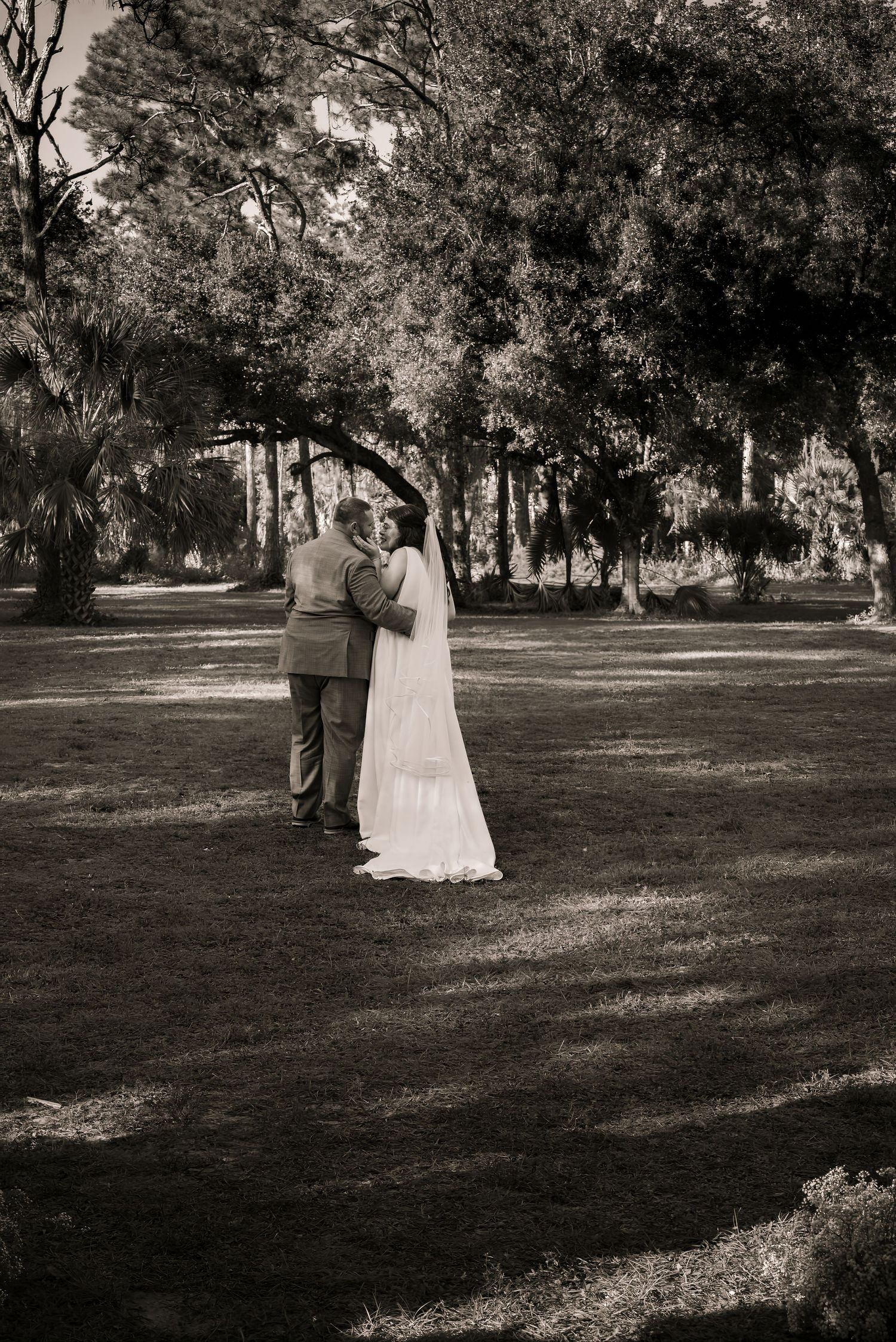 Black and white photo of bride and groom embracing under large trees in a sunlit garden.