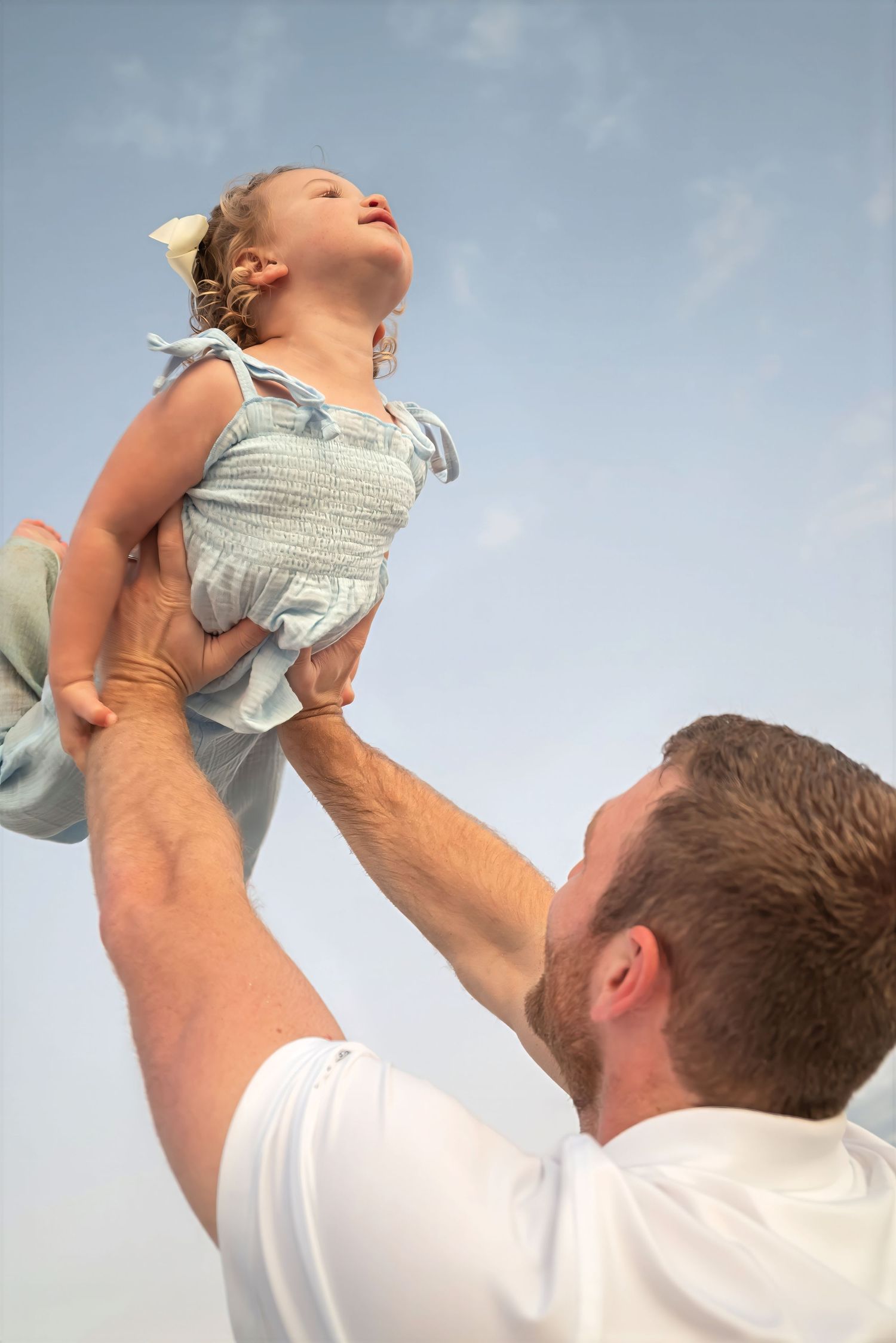 Father lifting laughing toddler girl in blue smocked dress high into a clear blue sky at the beach.
