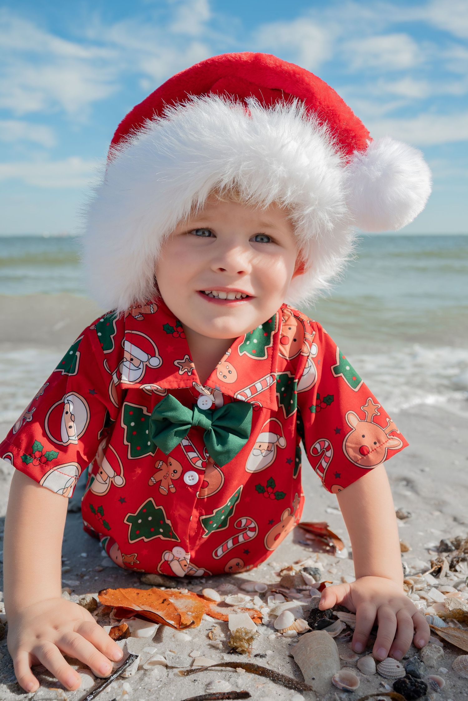Toddler in Santa hat and Christmas shirt playing on a sandy beach.