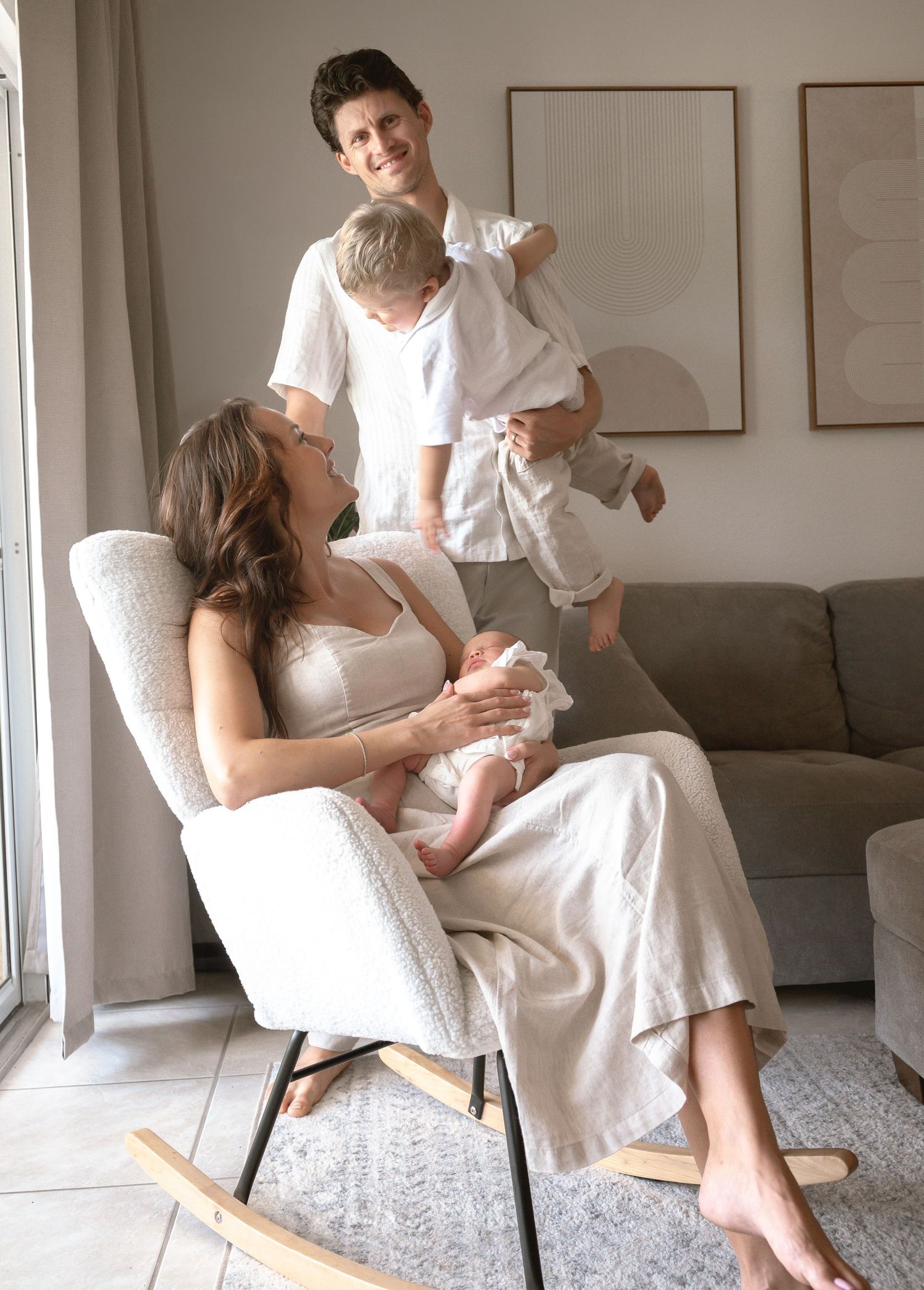 Happy family with mother holding newborn in rocking chair while father holds toddler nearby.