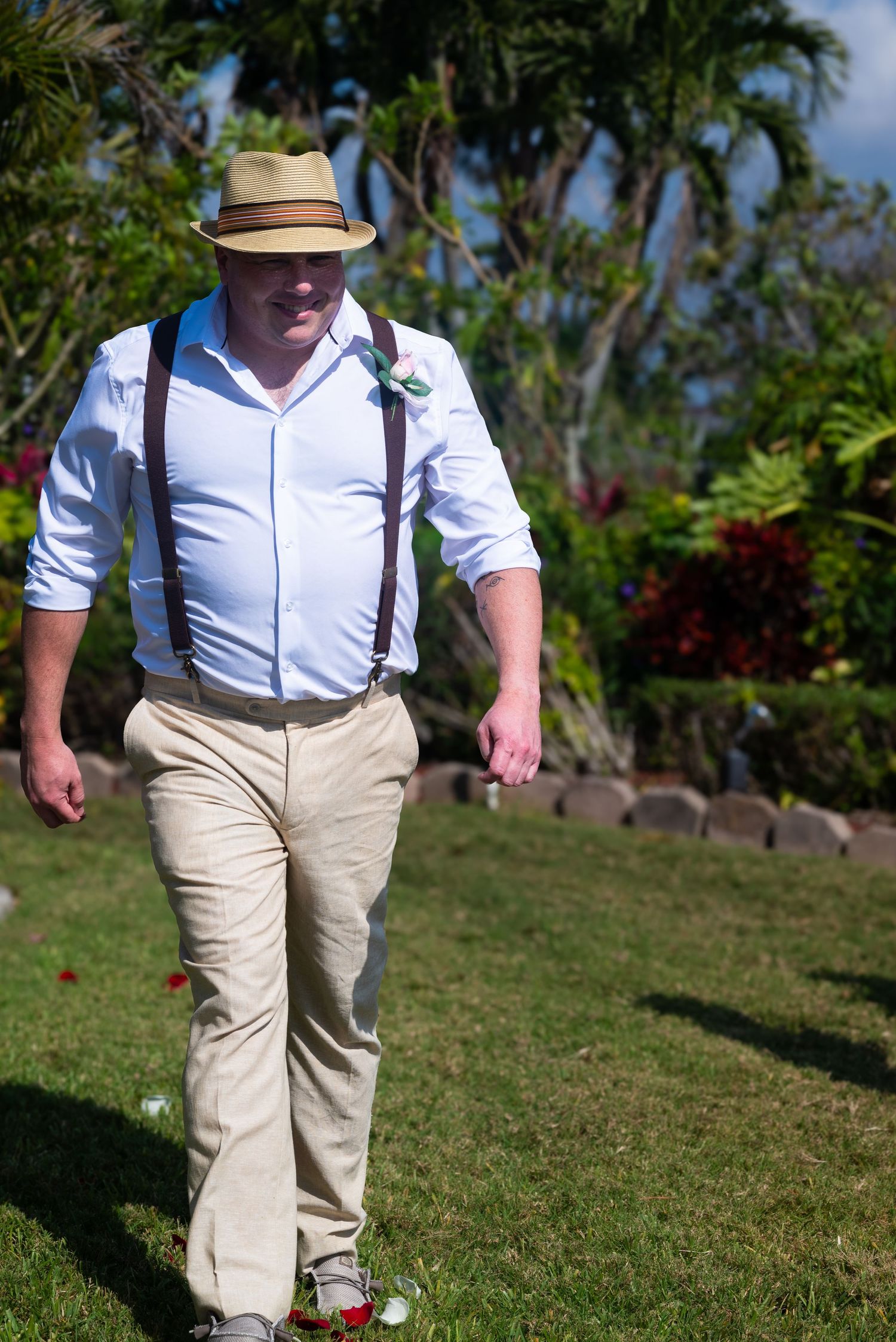 Groom in straw hat, white shirt, suspenders, and khaki pants walks across green lawn at outdoor wedding.