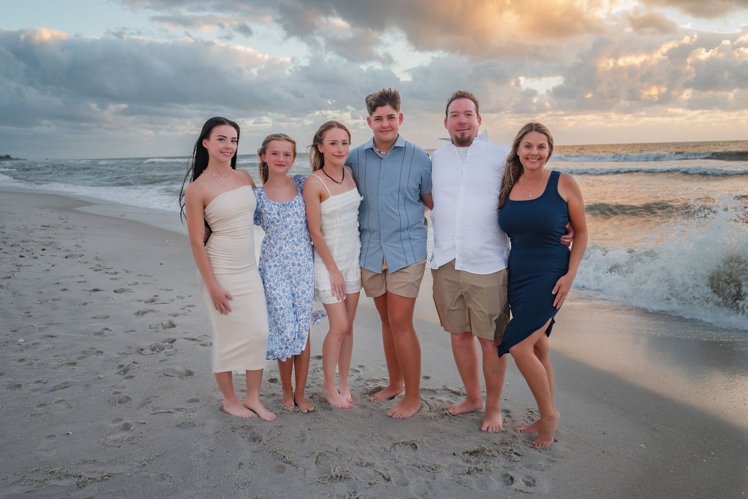 Family of six poses together on a sandy beach with dramatic sunset clouds in the background.