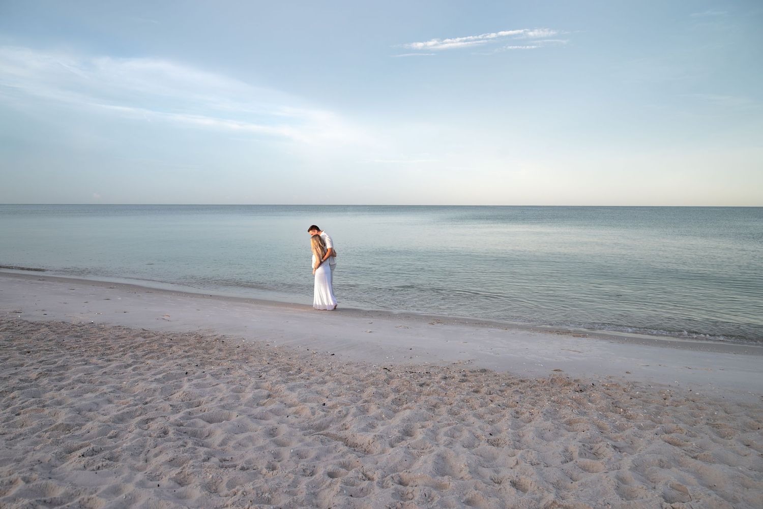 Pregnant couple walking along a serene white sand beach at dusk with calm turquoise ocean in background.