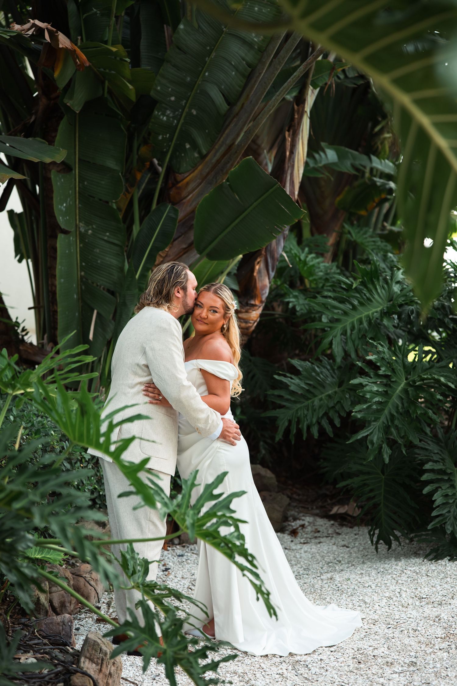 Bride and groom embracing among lush tropical greenery, bride in off-shoulder white gown, groom in cream suit.