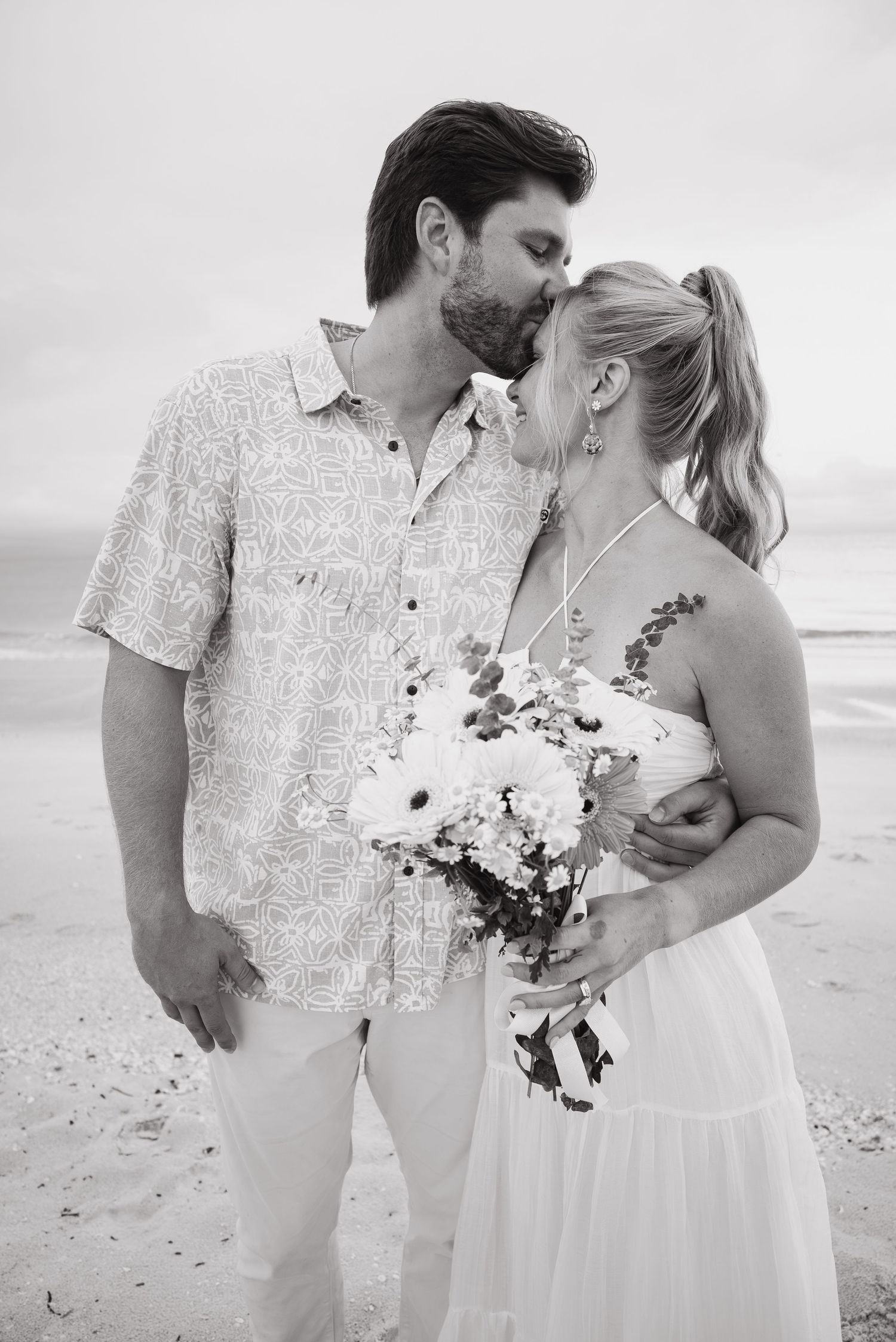 Black and white beach wedding photo of a couple kissing, bride holding a floral bouquet, ocean in the background.