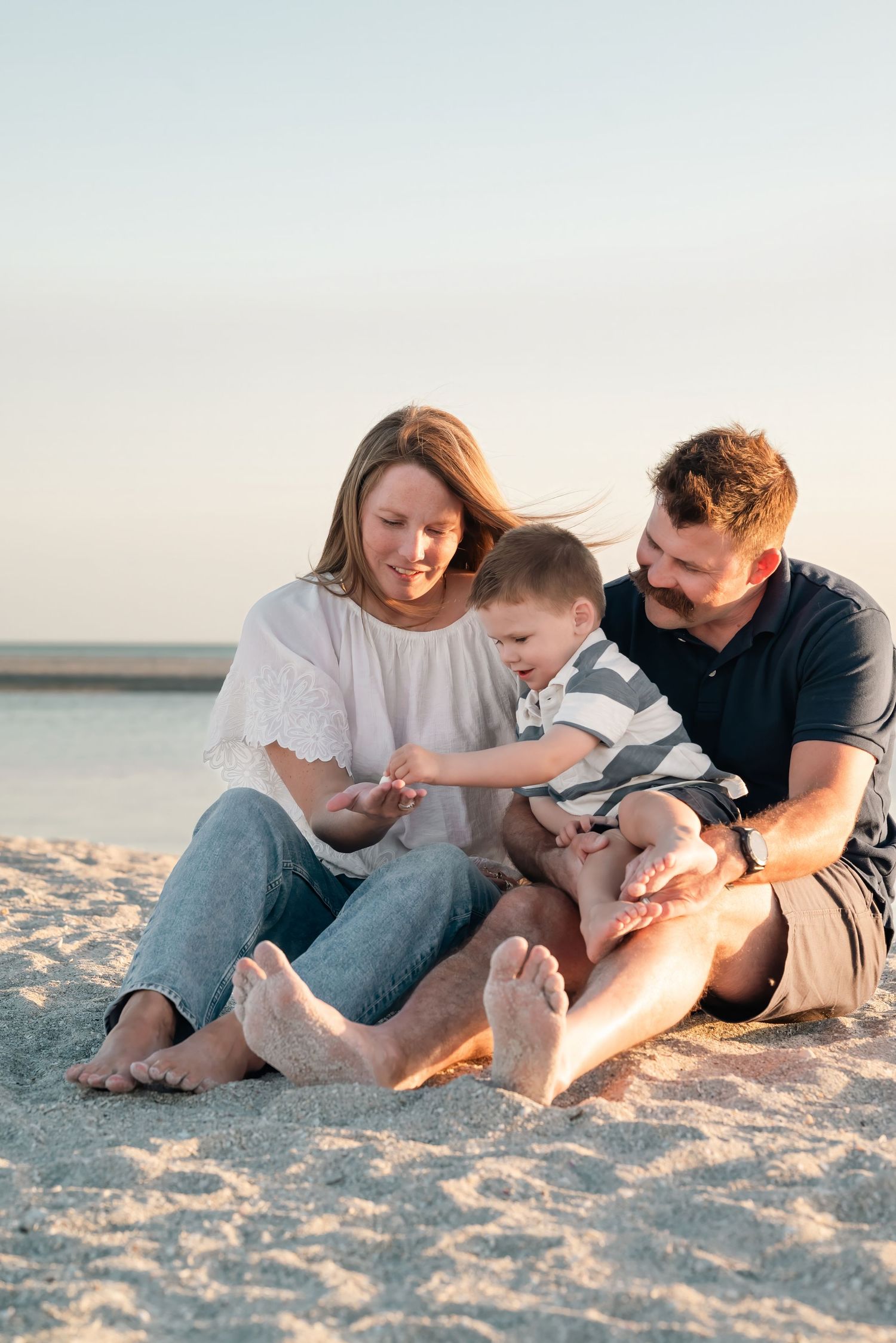 A happy family of three sits barefoot on a sandy beach at sunset, with parents smiling at their young toddler son.